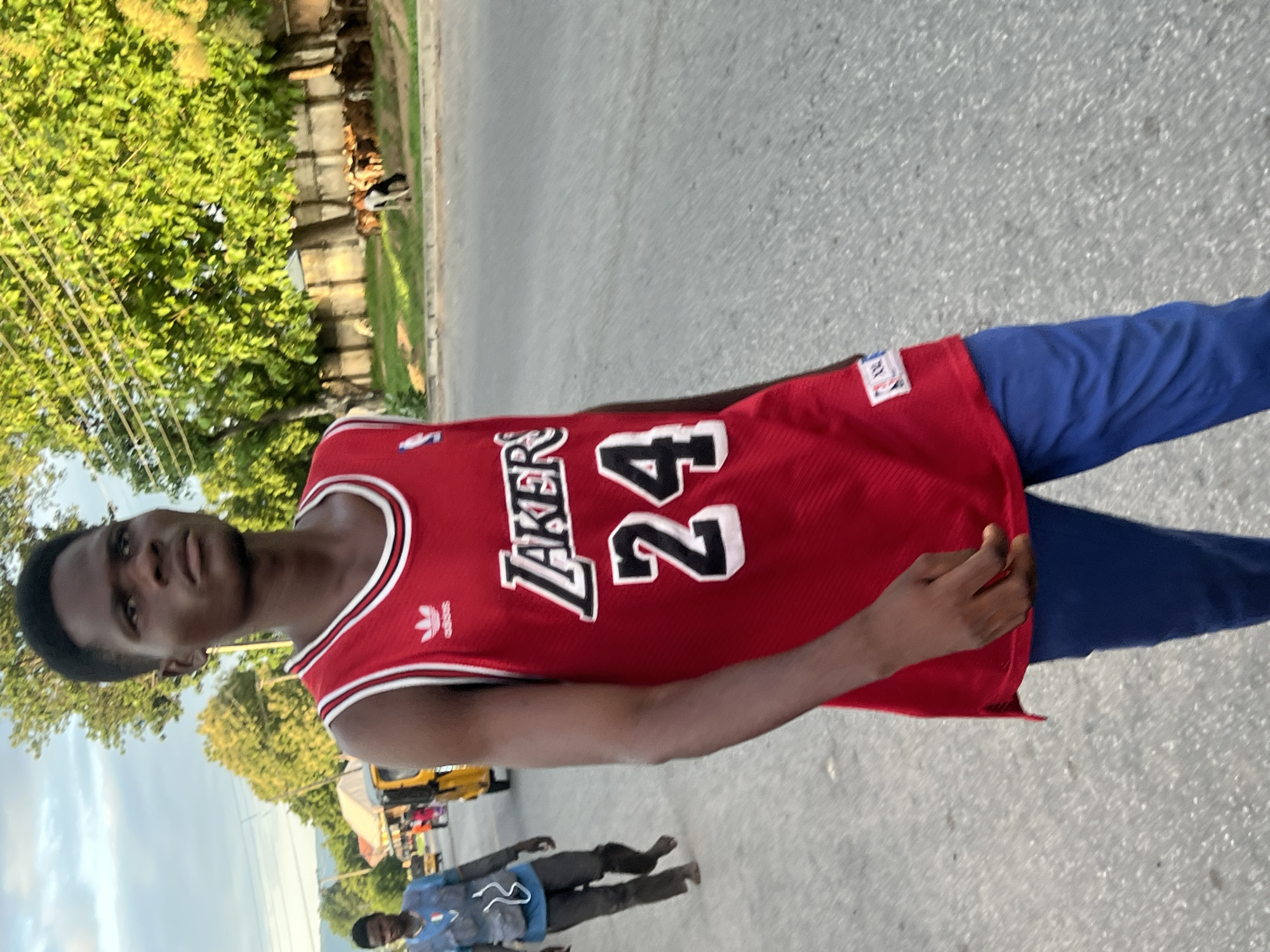 The image depicts a street scene in Jalingo, Nigeria. In the foreground, a dark-skinned young man walks forward and slightly to the viewer's right. He is wearing a red sleeveless basketball jersey with "LAKERS" and the number "24" in white with black outlining on the chest, and a white Adidas logo above. He is also wearing royal blue trousers. His expression is neutral, and his gaze is directed towards the right.
In the mid-ground on the left, another dark-skinned man walks, partially facing away from the viewer. He is wearing a light blue top with white detailing and dark pants, and appears to be barefoot. Behind him, a yellow auto-rickshaw (keke Napep) is partially visible, indicating active street traffic.
The background on the right features a grassy area with numerous green trees behind a low wall. Several goats and what appear to be cattle are visible grazing in this area. The sky above is light blue with scattered white clouds, suggesting daylight conditions. The ground surface is asphalt pavement.