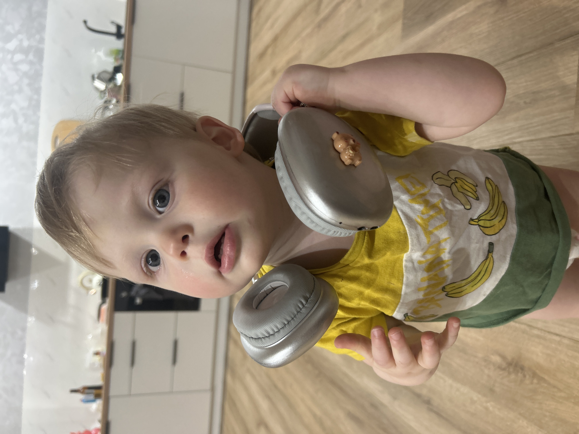 A young child, likely a toddler, is depicted in a close-up, chest-up shot within an indoor setting. The child has light blonde hair and bright blue eyes, looking directly at the camera with a curious, slightly open-mouthed expression and rosy cheeks. They are wearing a vibrant yellow and white t-shirt featuring yellow banana illustrations and the partial text "BANANAS" across the front, paired with green shorts or a diaper cover.
Large, silver-grey over-ear headphones are draped around the child's neck and shoulders. A small, light brown, possibly teddy bear-shaped charm is conspicuously attached to the outer shell of the left earcup. The child's small hands are visible, one lightly gripping the headphones.
The background reveals a modern kitchen area in Chaikovsky, Russia. Light wooden laminate flooring is visible in the foreground. Behind the child, white kitchen cabinets with dark handles can be seen, along with a glimpse of a wall with a subtle pattern and what appears to be a dark faucet or fixture. The lighting suggests it is daytime.