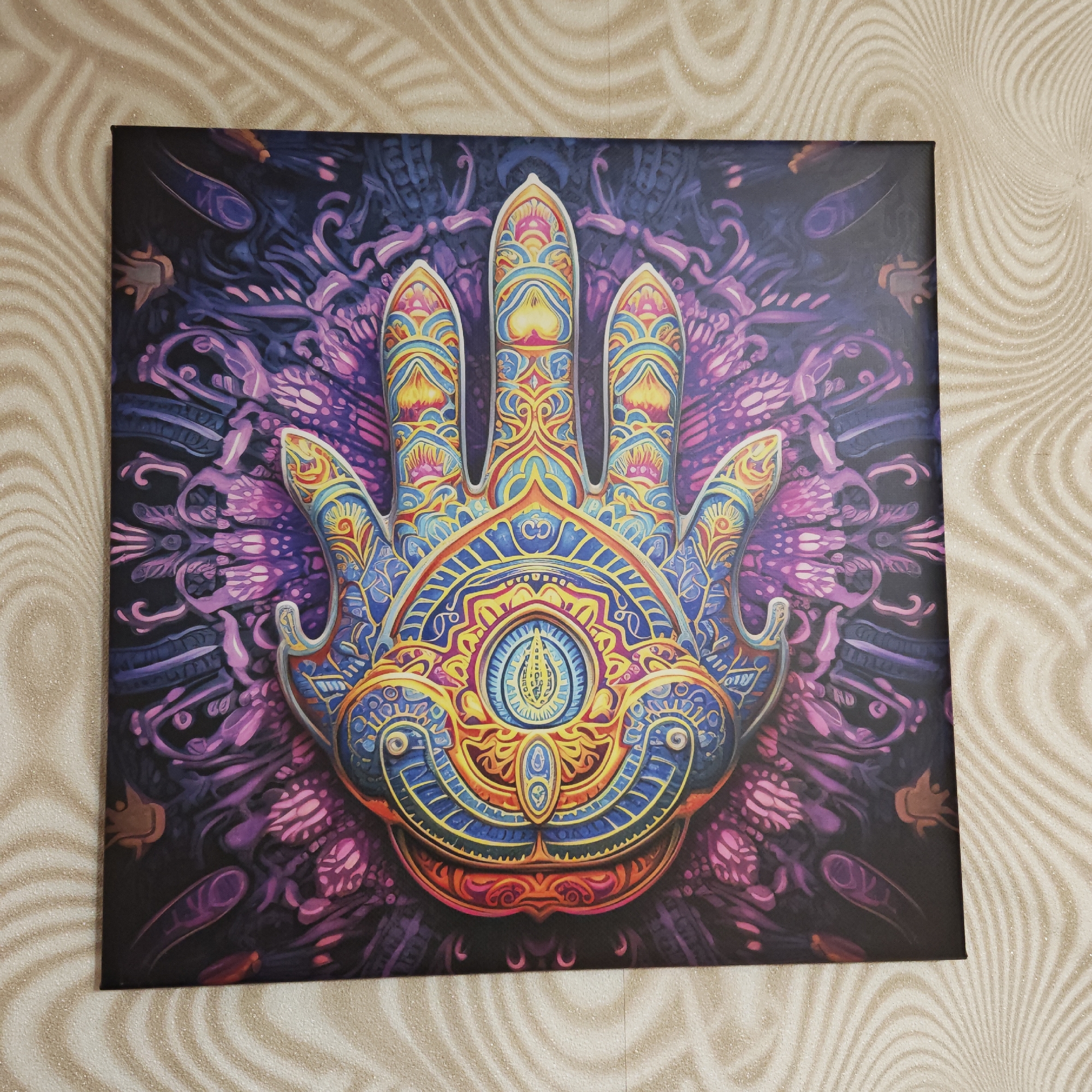 A vibrant, stylized artwork of a Hamsa hand, likely a canvas print, is prominently displayed against a patterned wall. The central subject is an ornate hand, rich in detail and color, dominating the frame. It features an intricate design with a palette of blues, yellows, reds, oranges, purples, and pinks. The palm of the hand holds a distinctive, elongated "eye" motif, above which a clear "Om" symbol is visible. The fingers and the surrounding areas of the hand are adorned with elaborate, symmetrical patterns and flowing designs, characteristic of spiritual or psychedelic art. The artwork's background is dark, primarily deep blue and purple, with swirling, organic patterns in lighter purple and pink tones, reminiscent of mandalas. The artwork is hanging indoors on a wall covered with a light-colored, textured wallpaper that exhibits a subtle, wavy, and swirling pattern in beige or cream. There is no specific activity taking place, as the image captures a static display of the art. The scene is located in Kladovitsy, Russia.
