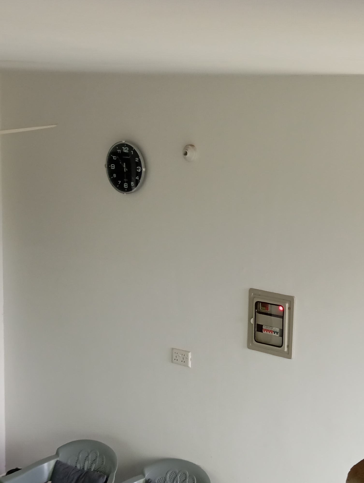 An indoor scene shows a white wall with a black circular clock. A small, white circular object is also mounted on the wall. Below this, a gray metal electrical panel is visible, with a red light illuminated inside. To the left of the electrical panel, a white electrical outlet is mounted on the wall. In the lower part of the image, two light gray chairs with dark cushions are partially visible. The room appears to be empty and still.