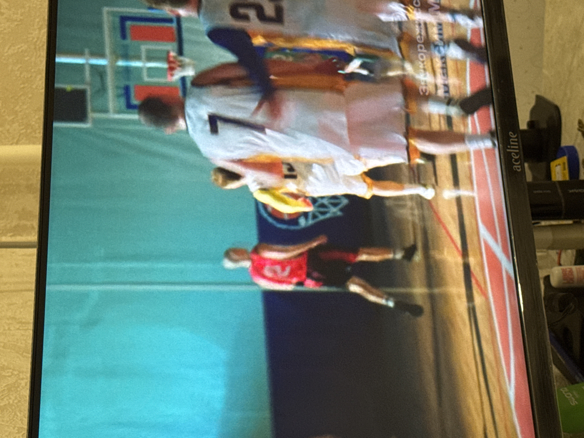 The image depicts a television screen displaying a basketball game. In the foreground, the lower part of a TV screen is visible with a brand name "aceline" on the bottom bezel. On the screen, several basketball players in uniform are engaged in the game. One player in a red jersey with the number 2 is positioned towards the left, facing away from the camera. To his right, a player in a white jersey with the number 7 is visible from the back. Another player in a white and yellow jersey is slightly to the left and behind the player with number 7. Further to the right, parts of other players in white and blue uniforms are visible. The court is wooden with red and white markings. The background shows a teal-colored wall and a basketball hoop with red and blue accents. Text is overlaid on the screen, partially obscured, appearing to be in Russian Cyrillic script. The overall image quality is somewhat blurry, indicating it was taken of a screen. The location is not discernible from the image itself, but it is stated to be in the city of Saratov, Russia.