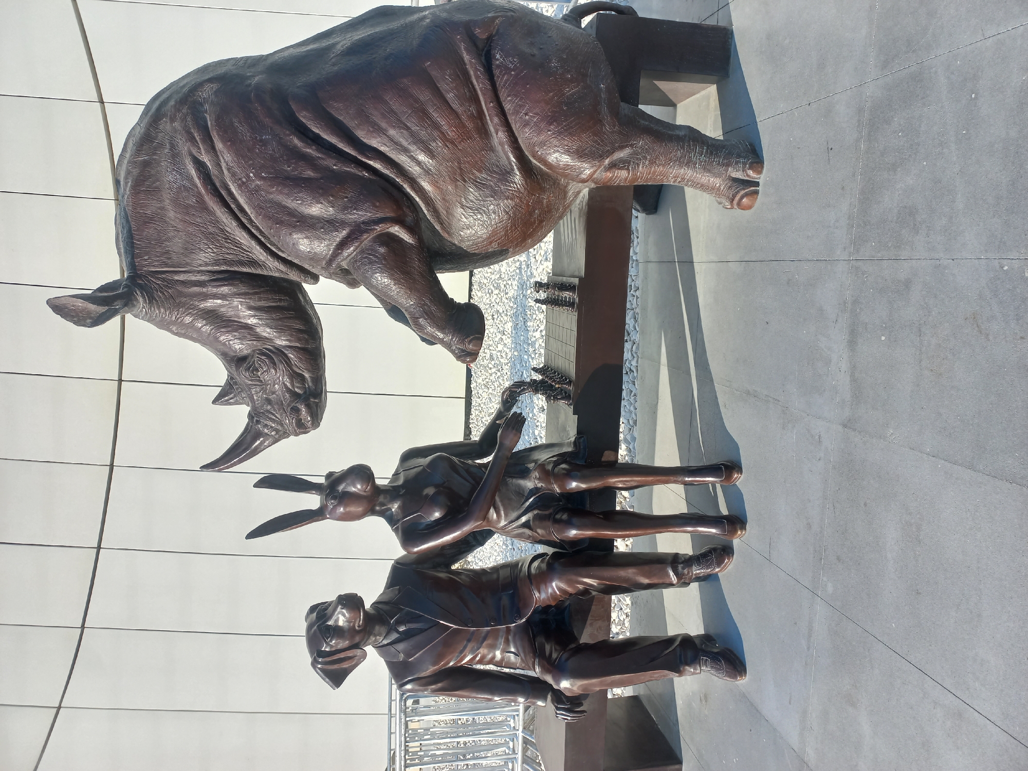 A bronze sculpture depicts three anthropomorphic animals playing chess on a bench in Baku, Azerbaijan. On the left, a dog dressed in a suit sits, leaning towards a rabbit in a dress who is seated beside him. The rabbit is reaching towards the chess board between them, while the dog appears to be observing the game. To the right of the bench, a large rhinoceros statue stands, its head turned towards the chess game. The sculptures are made of dark bronze and are set against a backdrop of a modern building with a white, paneled facade. The ground is paved with large, square grey tiles, and there are white gravel stones scattered around the base of the bench and the rhinoceros. A metal railing is visible in the background, on the left side of the image. The lighting suggests it is daytime, with distinct shadows cast by the sculptures.