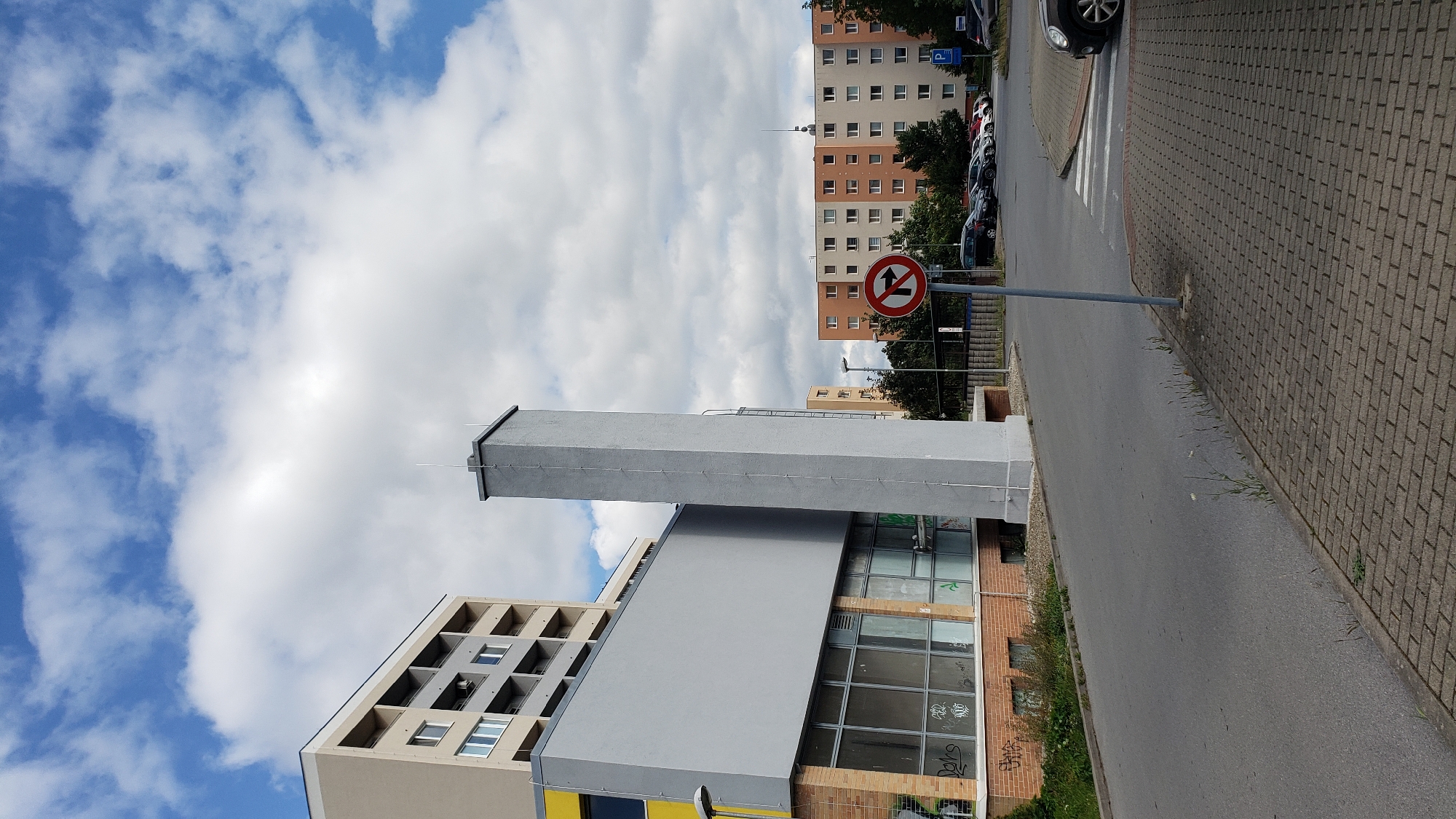 The image depicts an outdoor scene in the city of Prešov, Slovakia, featuring urban architecture and a street. In the foreground, a grey paved walkway is visible, leading to a paved street. On the right side of the street, a "no right turn" traffic sign is mounted on a pole. Beyond the street, a row of parked cars is visible, and further in the background are several multi-story apartment buildings. A tall, narrow, grey concrete structure, possibly a ventilation shaft or chimney, stands prominently in the mid-ground. Adjacent to it, a modern building with a grey facade and large windows, some exhibiting graffiti, is present. Another apartment building, with a beige and grey exterior, is seen to the left. The sky above is partly cloudy with cumulus clouds against a blue sky. The overall scene suggests a residential or mixed-use urban environment.