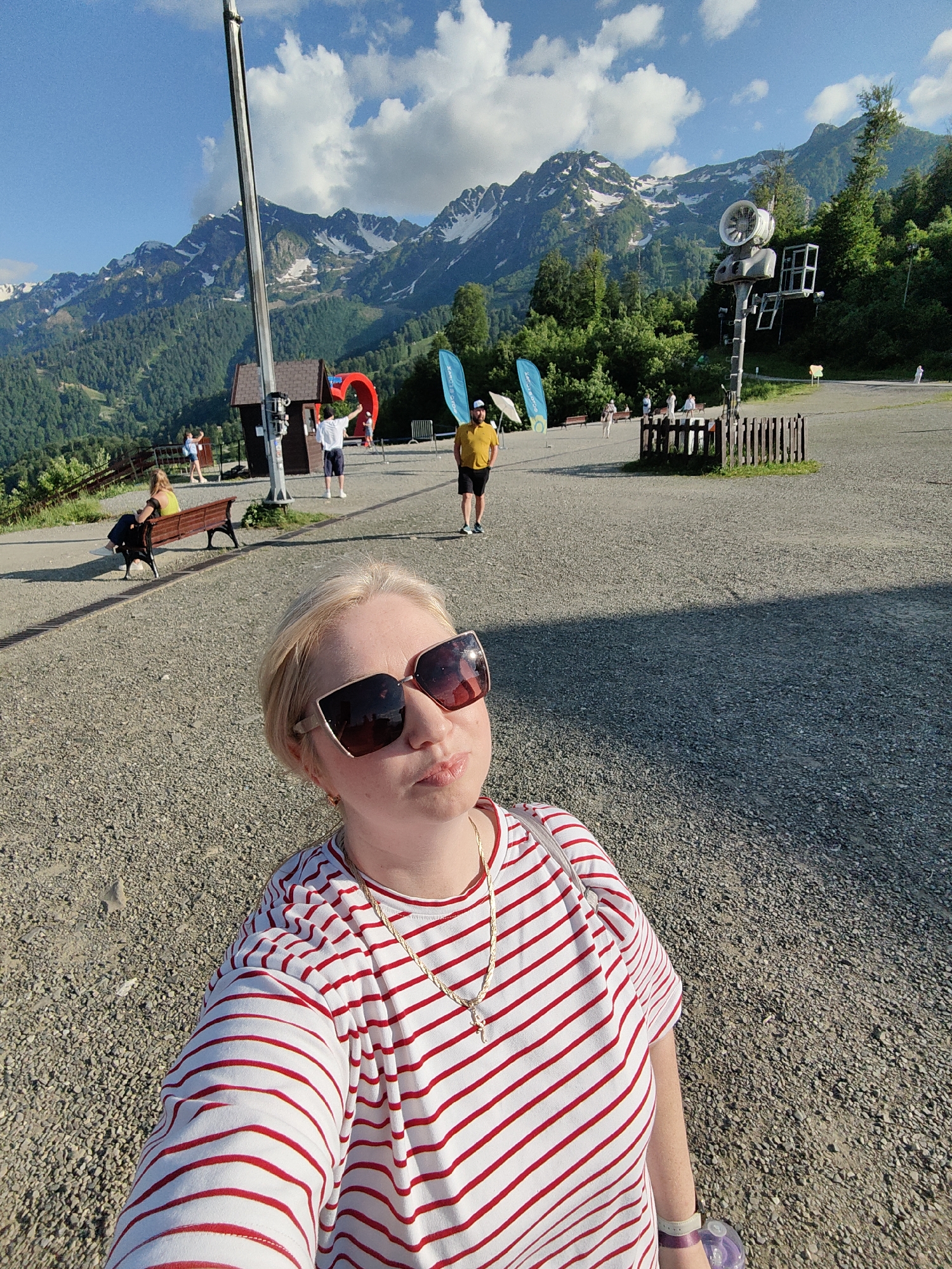 This is a medium shot, selfie-style photograph taken outdoors during the day. The main subject is a woman in the foreground, wearing a red and white striped t-shirt, large sunglasses, and a gold chain necklace. She is looking towards the camera with a slight pout.
In the background, there is a group of people scattered around a gravelly outdoor area. Some are walking, while others are sitting on a wooden bench. Behind them, there are mountains with snow-capped peaks, lush green trees, and a bright blue sky with scattered clouds. A tall pole with some equipment attached to it stands prominently on the right side of the frame. There are also some blue flags and a small, brown building with a red archway. The overall impression is a scenic, mountainous location, likely a resort or tourist area, on a sunny day. The woman's expression and the bright lighting suggest a pleasant, possibly vacation-like atmosphere.
There is no visible text in the image.