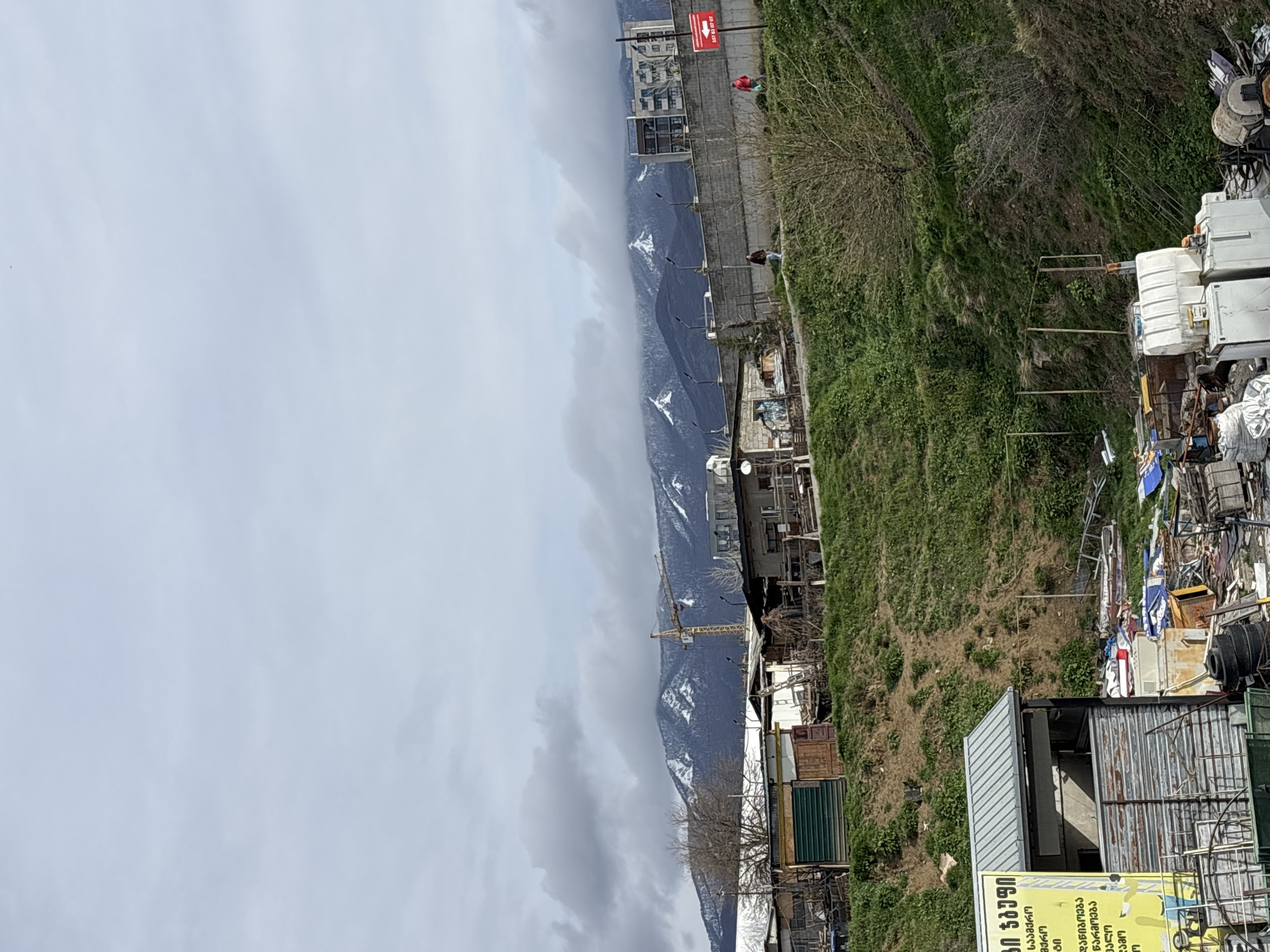 The image shows a mountainside in Tbilisi, Georgia, with snow visible on the peaks. A construction crane stands among buildings that appear to be homes or small businesses. The foreground includes an area with accumulated metal waste or salvaged materials. A sign with Georgian script is visible, situated near a metal structure. The sky is overcast. Two people can be seen walking along a paved area near a wall.