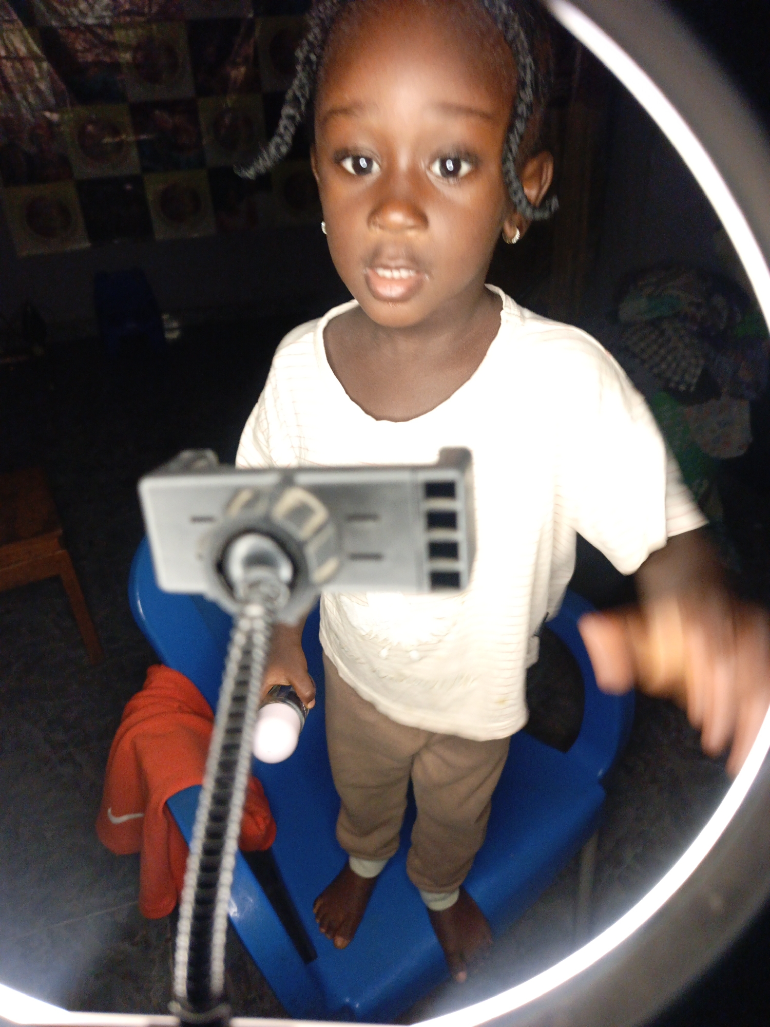 A young Black girl with braids and earrings is shown in a close-up shot. She is wearing a white t-shirt and brown pants. The girl is seated on a blue plastic chair. In front of her is a selfie ring light with a phone holder. A red jacket is draped over the chair. The background is dark and out of focus, with some patterned material visible. The lighting is strong, with the ring light illuminating the scene.