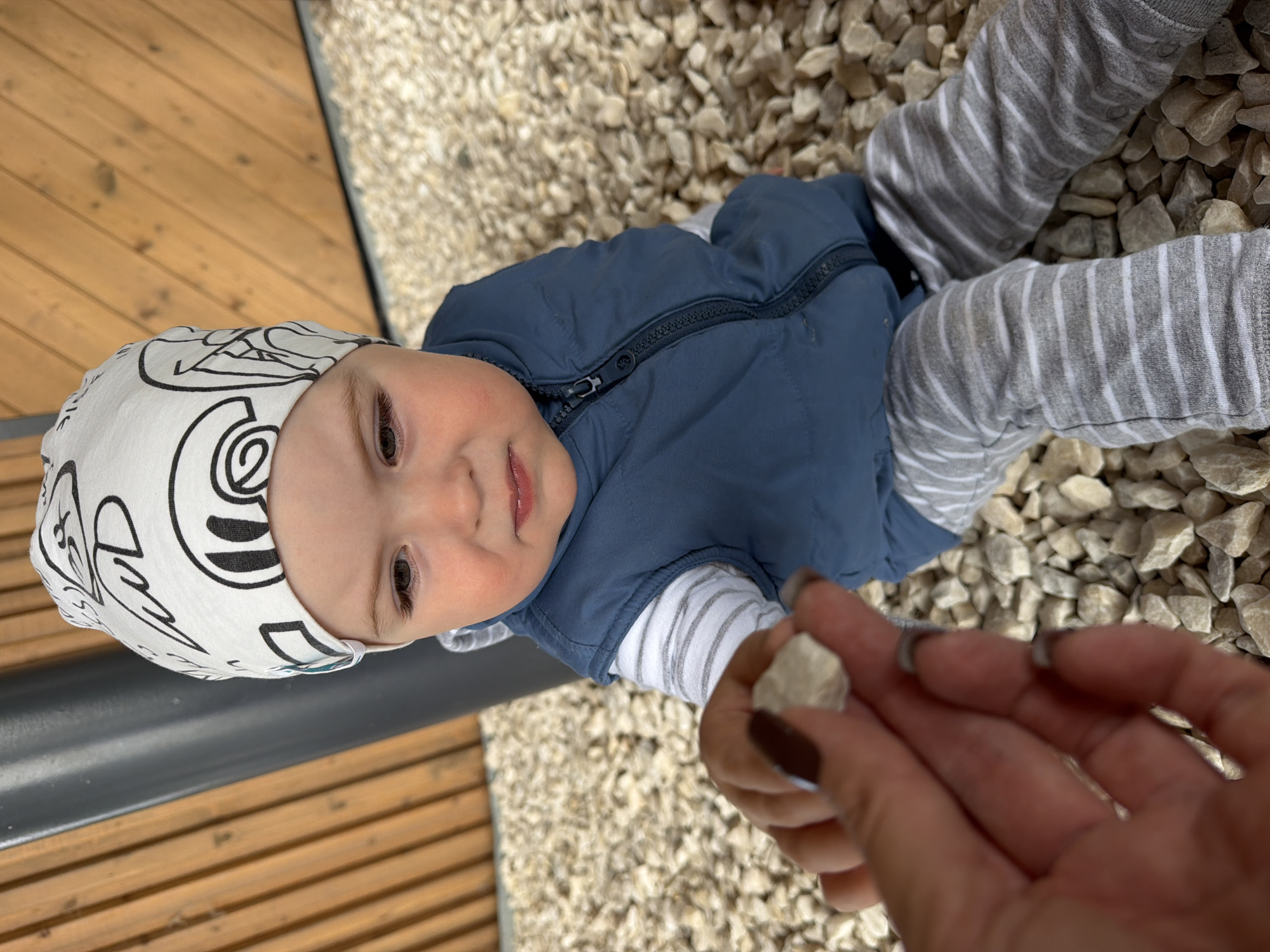 A close-up, high-angle shot shows a young child sitting on a bed of light-colored gravel. The child is wearing a white beanie with black line drawings, a blue vest over a striped long-sleeved shirt, and striped gray pants. The child's hands are clasped, and they appear to be looking at a small, light-colored pebble held in an adult's hand, which is in the foreground. The background consists of wooden planks and a dark gray pole. The scene is outdoors, likely in a park or yard. The location is Votkinsk, Russia.