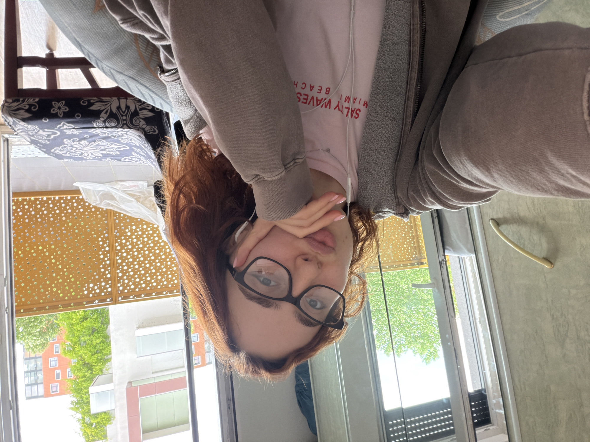 This upside-down selfie captures a young woman indoors, likely in an apartment in Strasbourg, France. She has reddish-brown hair, wears black-rimmed glasses, and has wired earphones in. Her left hand, adorned with light pink nail polish, partially covers her mouth and chin, with her tongue slightly visible in a playful or pouty expression.
She is dressed in a light pink t-shirt, on which the text "SALTY WAVES MIAMI BEACH" is visible (upside down), layered under a grey hooded zip-up jacket.
The background shows an outdoor urban environment through a large window or balcony door, illuminated by bright daylight. Green trees are visible, along with multi-story buildings featuring a mix of architectural styles, some with reddish-brown walls and numerous windows, and others with modern white and grey facades. A distinctive golden-brown perforated screen, possibly a balcony railing or decorative panel, is prominent above the outdoor view.
Indoors, parts of a room are visible, including patterned fabric on what appears to be a chair or cushion (black with white floral designs) and a light blue patterned throw. The scene suggests a casual, relaxed moment inside, with a clear view of a bright, daytime urban landscape.