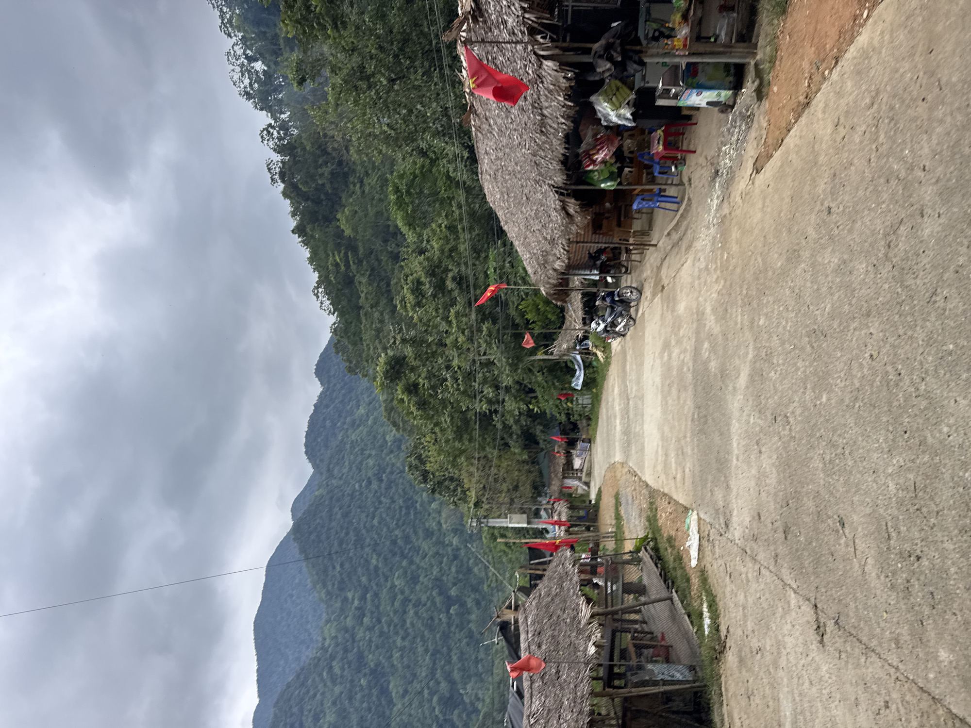 The image depicts a scene on a paved road in Cổ Lũng Commune, Vietnam, likely a rural area due to the surrounding lush green mountains. The sky is overcast with dense clouds. On both sides of the road, there are small shops with thatched roofs. Red flags with yellow stars, the national flag of Vietnam, are prominently displayed, some on poles and others attached to the structures. Several motorcycles are parked along the roadside in front of the shops. The shops appear to be selling various goods, with items visible on shelves and tables outside. The overall atmosphere suggests a quiet, perhaps slightly remote, community in a mountainous region.