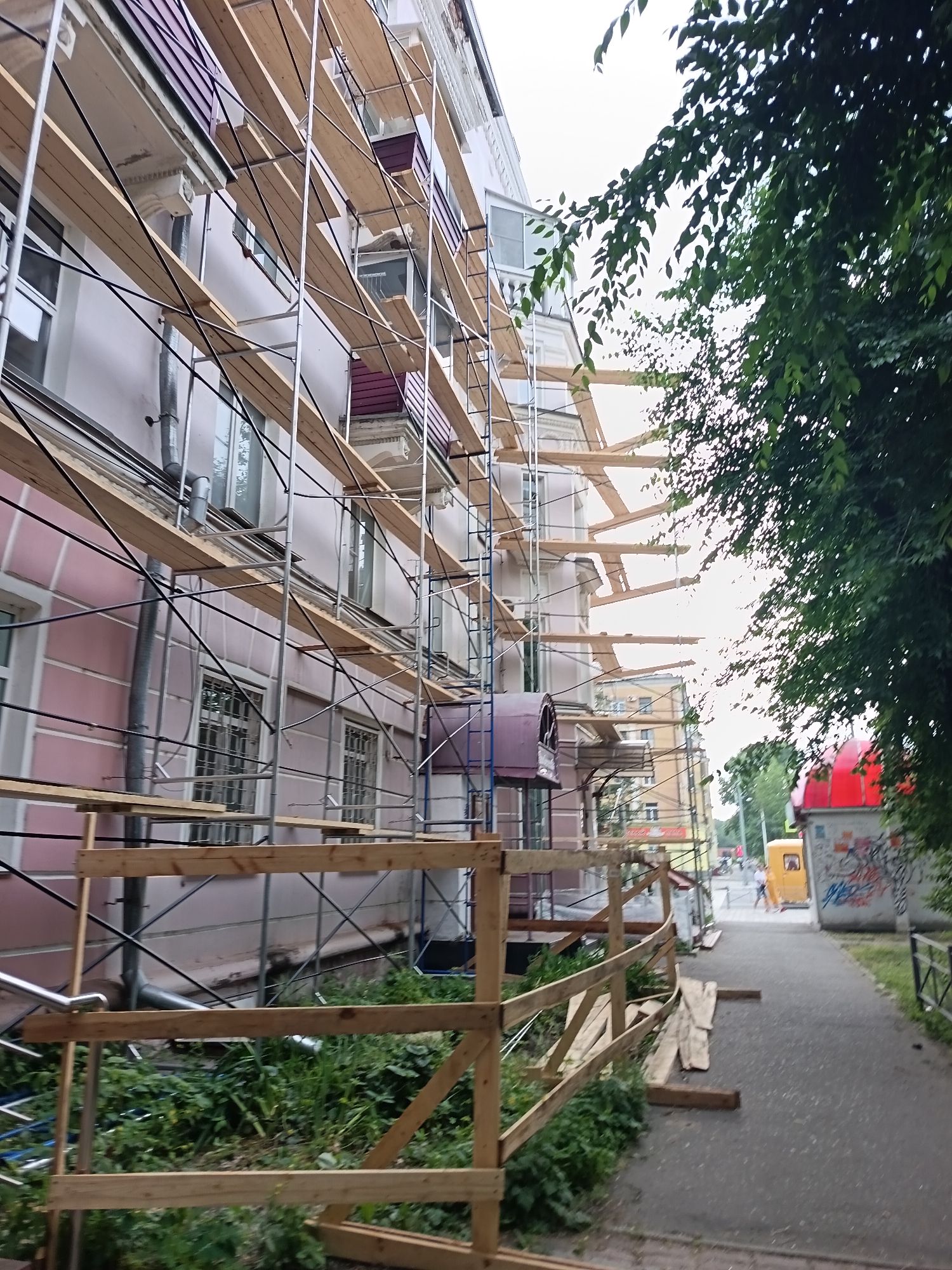 The image shows a section of a multi-story building undergoing renovation in Komsomolsk-on-Amur, Russia. The building's facade is painted a pale pink color and is covered in scaffolding made of metal poles and wooden planks. The scaffolding extends up the side of the building, obscuring many of the windows, some of which have metal grates. A drainpipe runs vertically along the facade. In the foreground, a temporary wooden fence has been erected, and various wooden planks are scattered around. To the right of the building, a paved walkway runs alongside it, leading towards a street scene. A yellow vehicle and a building with a red roof are visible in the background. Green foliage from trees overhangs the scene from the right side of the image. The sky is overcast, suggesting cloudy weather. There are no people clearly visible in the immediate foreground or interacting with the structure.