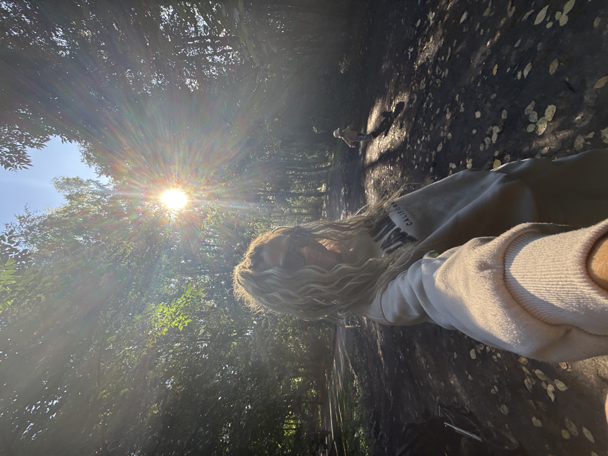 The image depicts a woman with long, blonde, curly hair, wearing sunglasses and a light-colored hooded sweatshirt, taking a selfie in a wooded area. The sun is low in the sky, creating strong lens flare and casting long shadows. In the background, a small child wearing a light-colored hat and jacket is walking away from the camera along a dirt path. The path is littered with fallen leaves. The setting appears to be a park or forest. The location is identified as the city of Rusanovo, Russia, although this cannot be visually confirmed from the image itself.