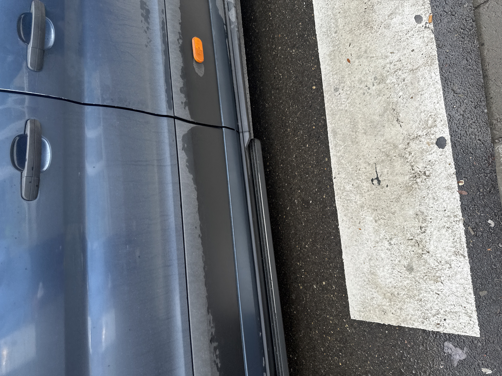 The image shows the side of a blue van parked on a wet asphalt road. The van has two doors visible, with black door handles. There is an orange reflector on the lower part of the van. Below the van's doors, a black side skirt is visible. In the foreground, a portion of a pedestrian crossing, marked by white and black stripes, is visible. The road surface is wet, suggesting recent rain. The scene appears to be on a street in Tbilisi, Georgia, based on the context of the image, though no specific landmarks are visible to confirm the location. There are no people or animals in the image. The overall condition of the van suggests it is in use, with some dirt and water marks on the body.