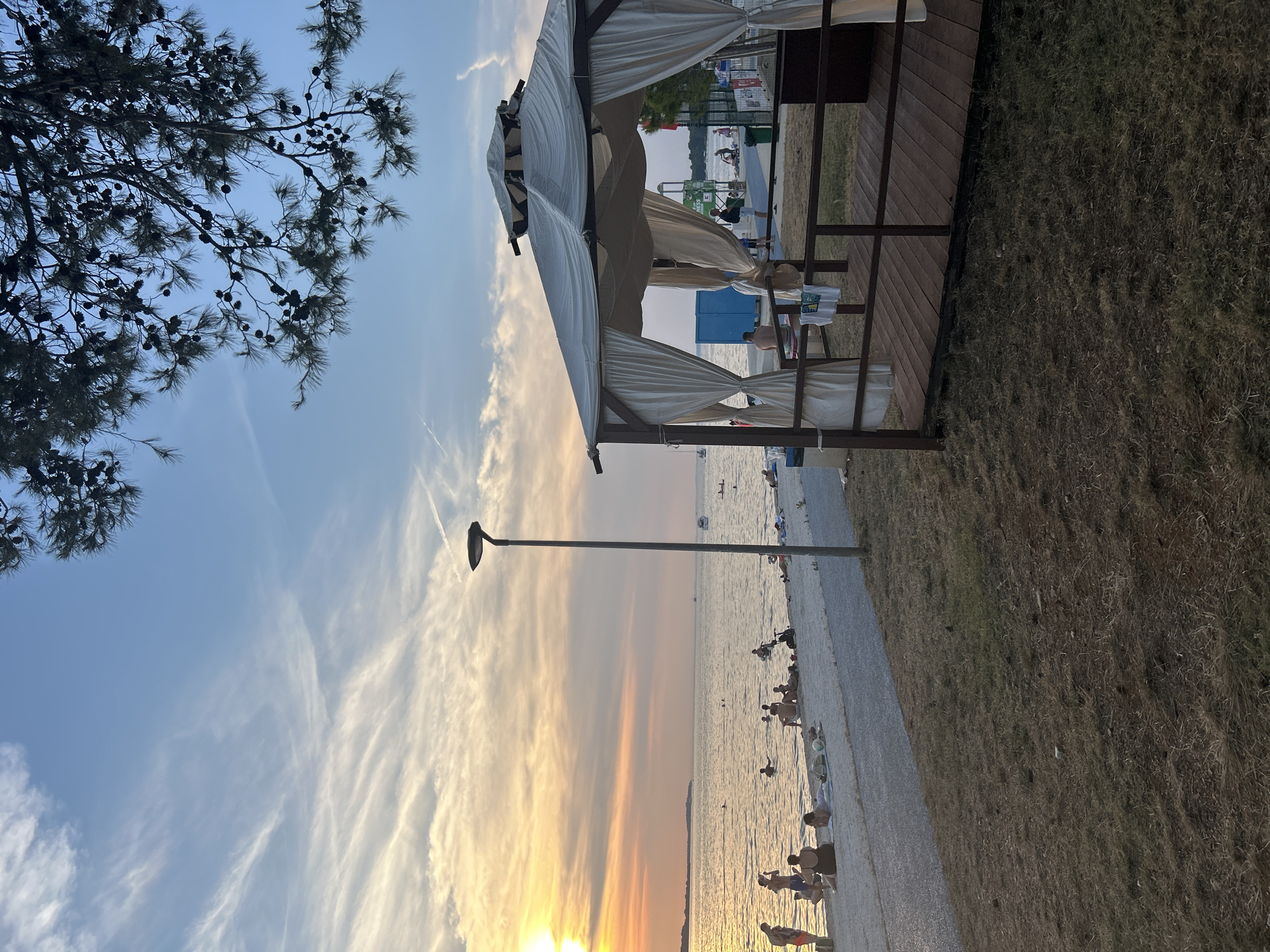 This is a landscape orientation, eye-level, full shot of a beach scene during sunset. In the foreground to the right, there is a wooden gazebo with white curtains that are pulled back. The gazebo has a wooden deck and railing. To the left of the gazebo is a dry, grassy area. Behind the gazebo and grass is a paved path. Beyond the path is a sandy beach where several people are gathered, some in the water and some sunbathing. The sea stretches out to the horizon, with a gentle sunset casting warm colors across the sky and water. A single lamppost stands on the path between the gazebo and the beach. In the distance, there are a few boats on the water and trees along the coastline. The sky is a mix of blue with wispy white clouds, tinged with orange and yellow from the setting sun. A tree with pinecones hangs from the top right corner of the image.