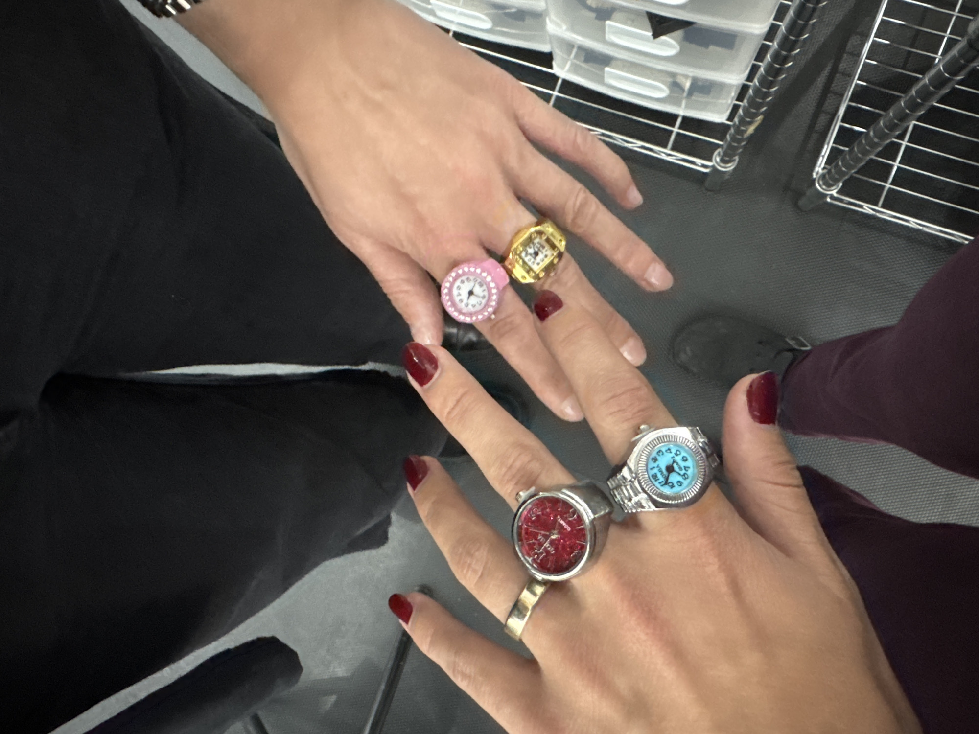 Two people display their hands adorned with novelty rings shaped like wristwatches. The person on the left wears black pants and has rings with a red face and a blue face, while the person on the right wears maroon pants and has rings with a pink face and a gold face. The background shows parts of metal shelving with plastic storage bins on top, and a dark gray floor. The location appears to be indoors, though the city, Mexico City, Mexico, cannot be confirmed from the image alone.