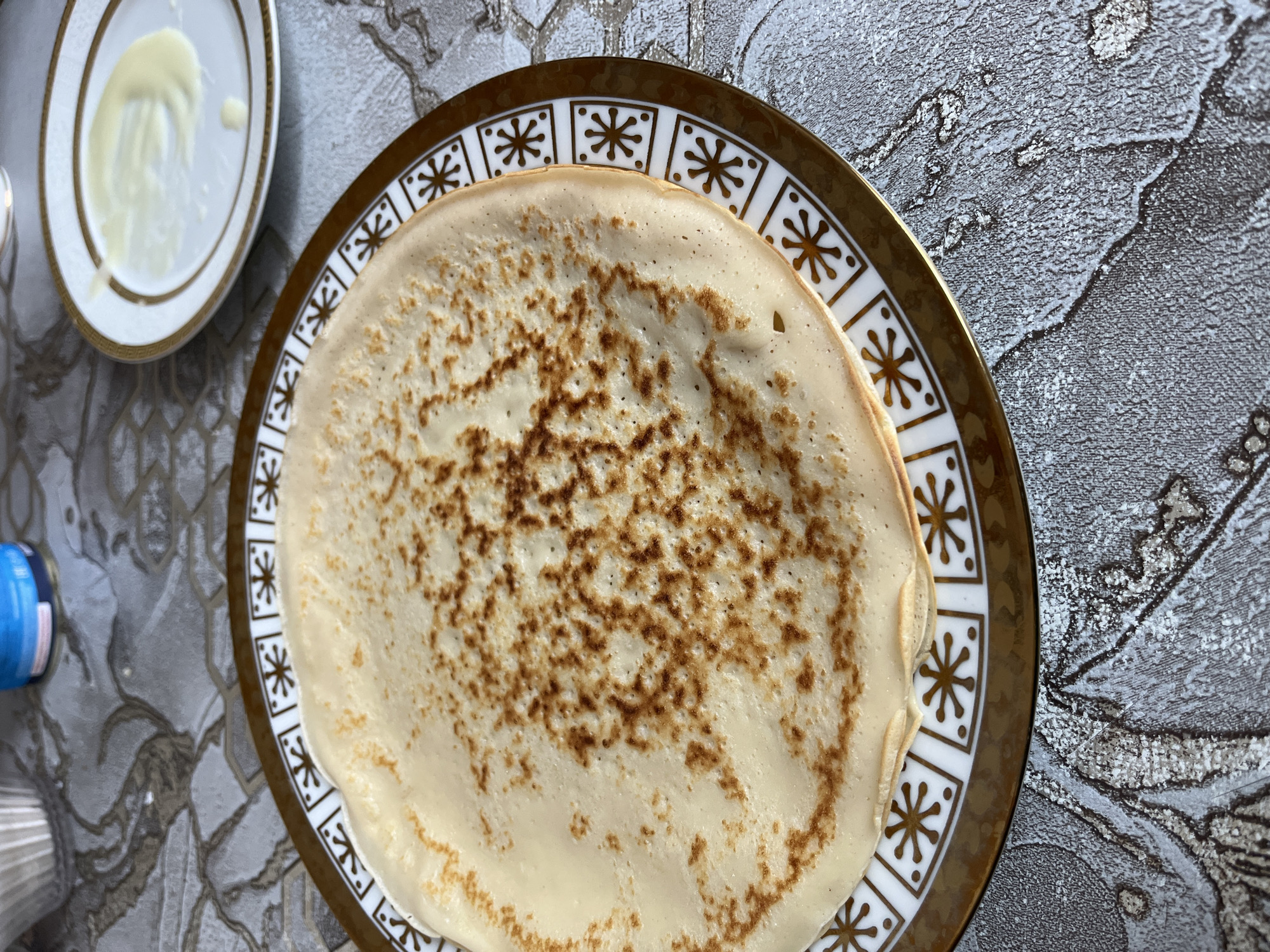 The image depicts a stack of pancakes on a decorative plate with a white and gold rim. The pancakes have a light golden-brown mottled pattern on the surface. A small plate with a dollop of white sauce is visible in the upper left corner. The plates are set on a surface with a grey and silver decorative pattern. The location is unspecified, but the user has indicated it is Kaspiysk, Russia.