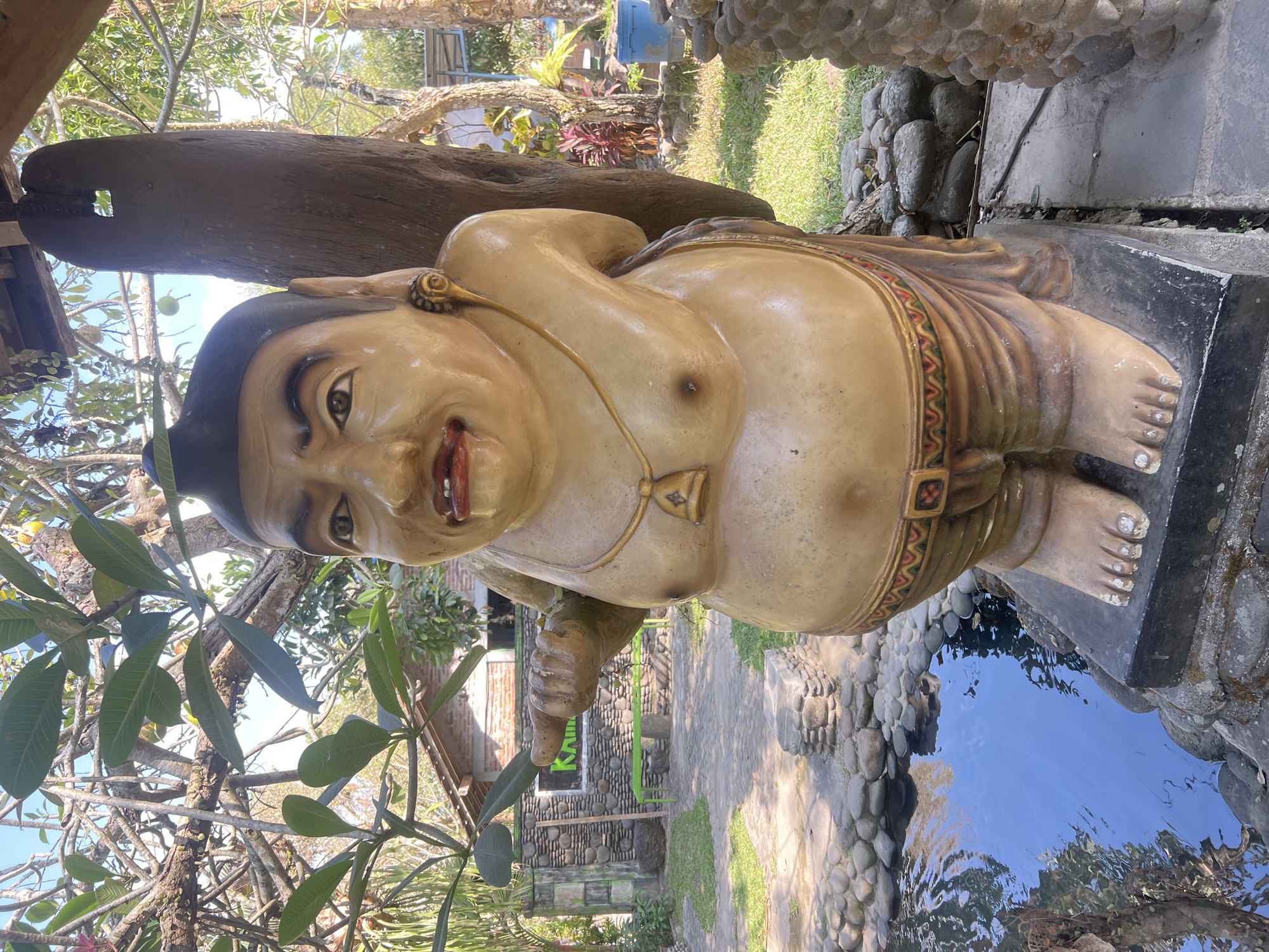 A weathered, tan-colored statue of a stout figure stands outdoors in a garden setting. The statue depicts a male with a wide belly, wearing a gold necklace with a bell pendant, and draped cloth around its waist and legs. Its skin is textured and shows signs of age. The figure has a large head with a stern expression, dark eyebrows, and exaggerated features, including a prominent nose and pursed lips. It is positioned on a dark grey rectangular base.
The statue is situated near a small, blue-tiled pond filled with clear water, surrounded by smooth, grey river stones. Beyond the pond, a paved area leads to a stone wall, behind which sits a rustic building with a red-tiled roof and a green bench. The area is lush with vegetation, including trees with broad leaves and grasses. Sunlight casts shadows across the scene, indicating it is daytime. The overall environment suggests a tranquil, outdoor recreational space, likely a garden or a resort, in Tlogoadi, Indonesia.