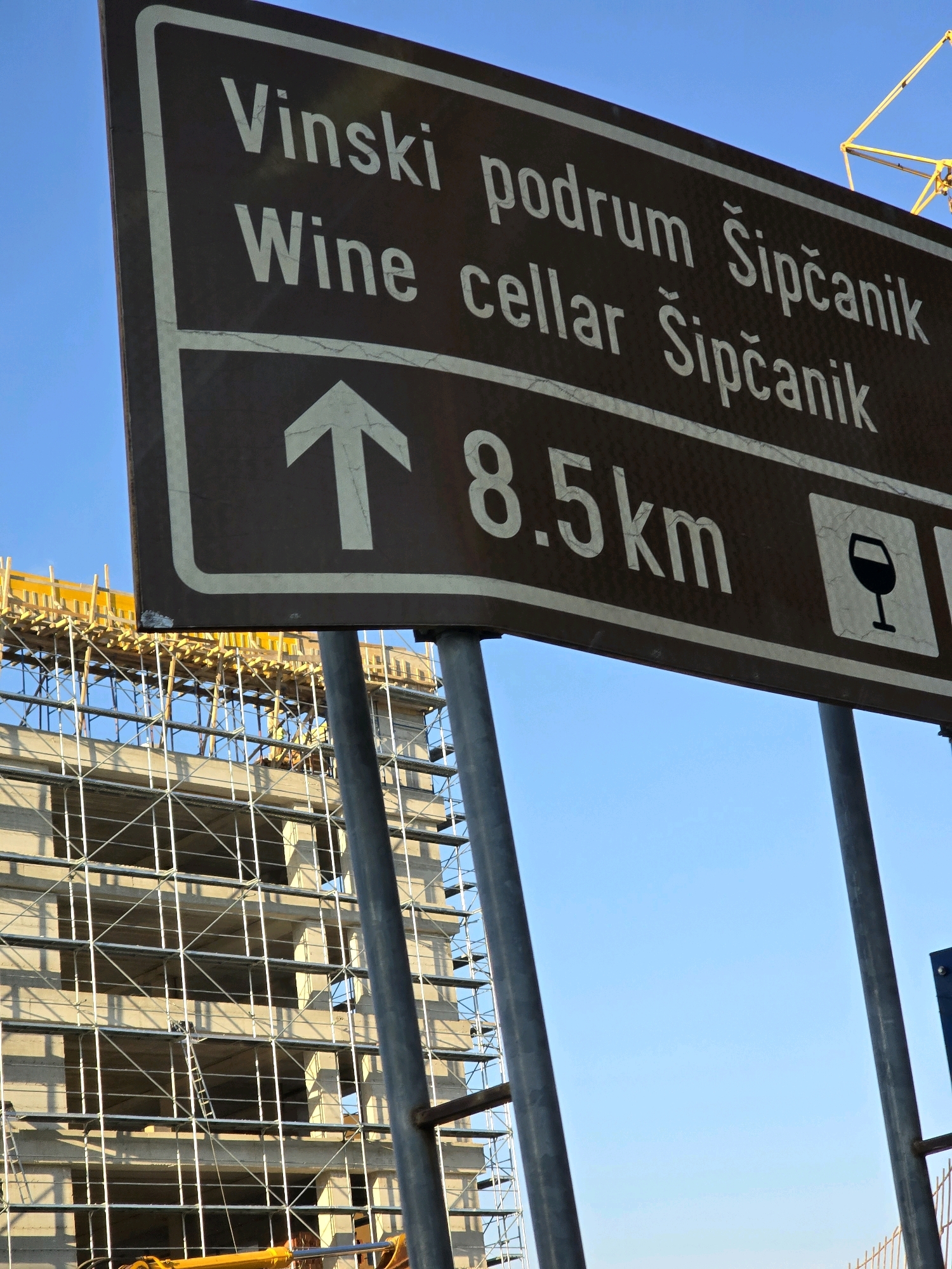 A brown directional sign with white text points the way to "Vinski podrum Šipčanik" and "Wine cellar Šipčanik" at a distance of 8.5 km, with an icon of a wine glass. The sign is mounted on metal poles. In the background, a large, unfinished concrete building is covered in scaffolding. A yellow construction crane is visible in the upper right corner. The sky is clear and blue. The scene appears to be outdoors, likely on the outskirts of Podgorica, Montenegro, given the reference to Šipčanik which is a notable wine cellar located near the city. There are no visible people in the image.
