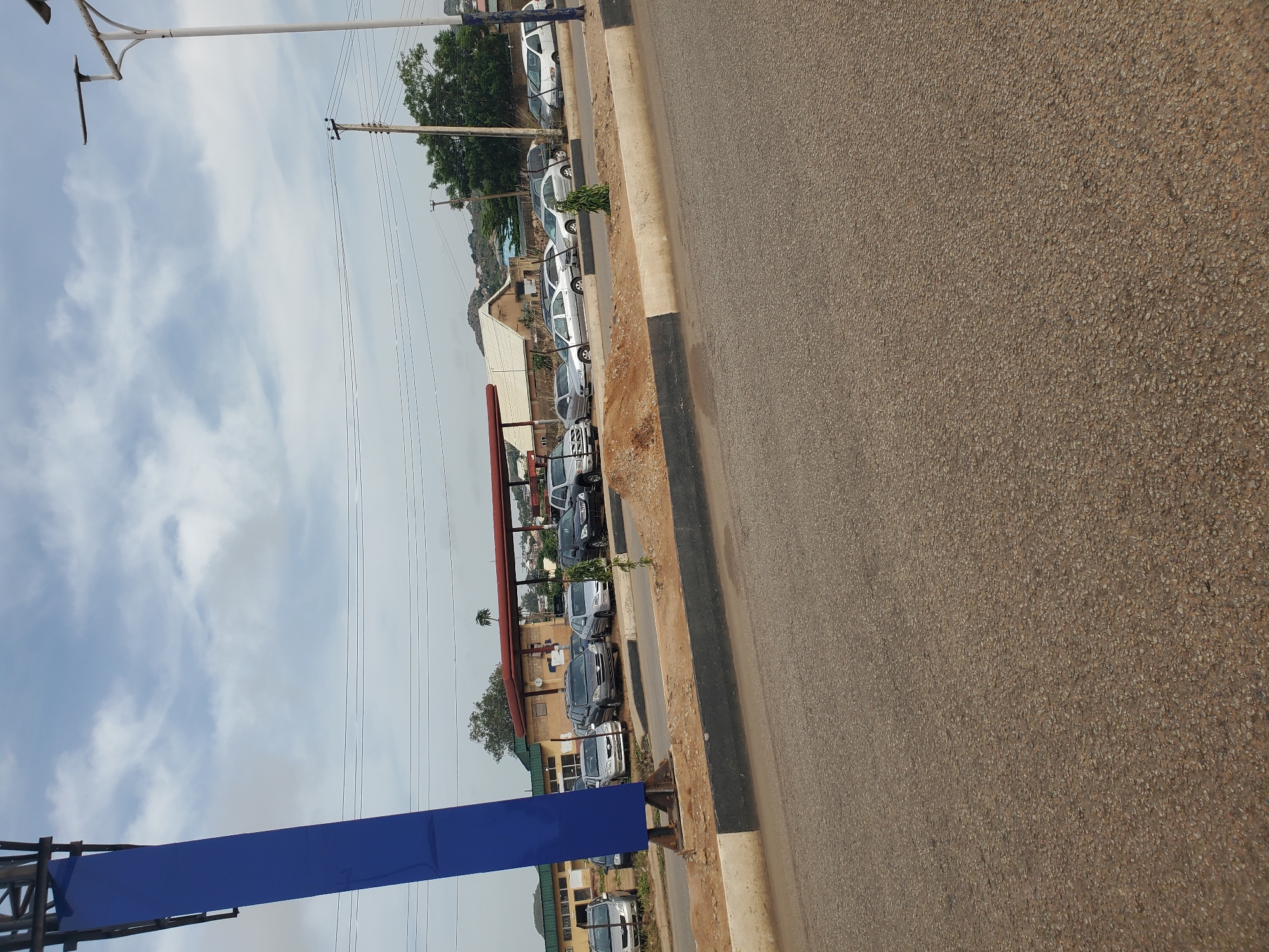 The image captures a daytime scene along a road in Bukuru, Nigeria, likely a commercial or dealership area given the presence of multiple vehicles parked. A prominent blue structure, possibly a sign or part of a building, dominates the left side of the frame. Several cars are visible parked in a lot to the right of the blue structure, suggesting a car sales lot or parking area. Beyond the parked cars, buildings with various roofing styles and colors are present, including what appears to be a residential area or more commercial establishments. The sky is overcast with scattered clouds, and utility poles with power lines are visible in the background, indicating an urban or semi-urban setting. The road in the foreground is paved and appears to be in regular use, with a concrete barrier separating it from the parking area. There are no people visible in the image, and the overall impression is one of a quiet, everyday scene in Bukuru.