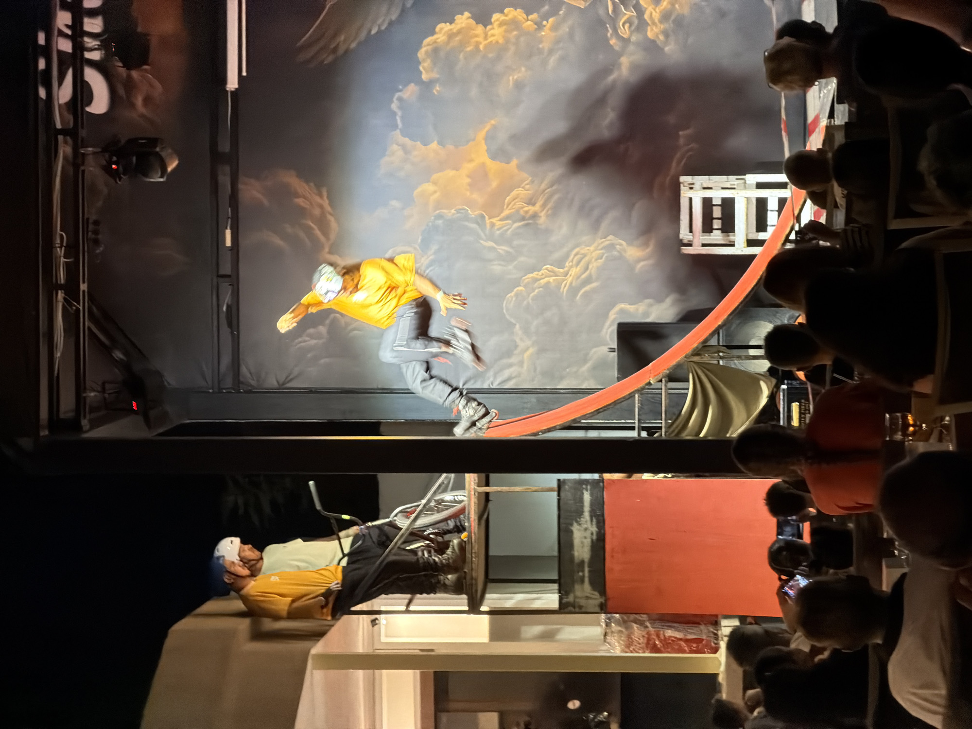 The image captures an outdoor nighttime performance in Manavgat, Turkey. On a raised platform, two individuals wearing helmets stand beside a BMX bike. To the right, an aerial performer in a yellow shirt is suspended mid-air, seemingly in the process of a stunt. The backdrop is a large mural depicting a cloudy sky with warm lighting, creating an illusion of depth. In the foreground, an audience is seated, facing the performance area. The scene suggests a live entertainment event, possibly a circus or a stunt show, taking place in a public venue.