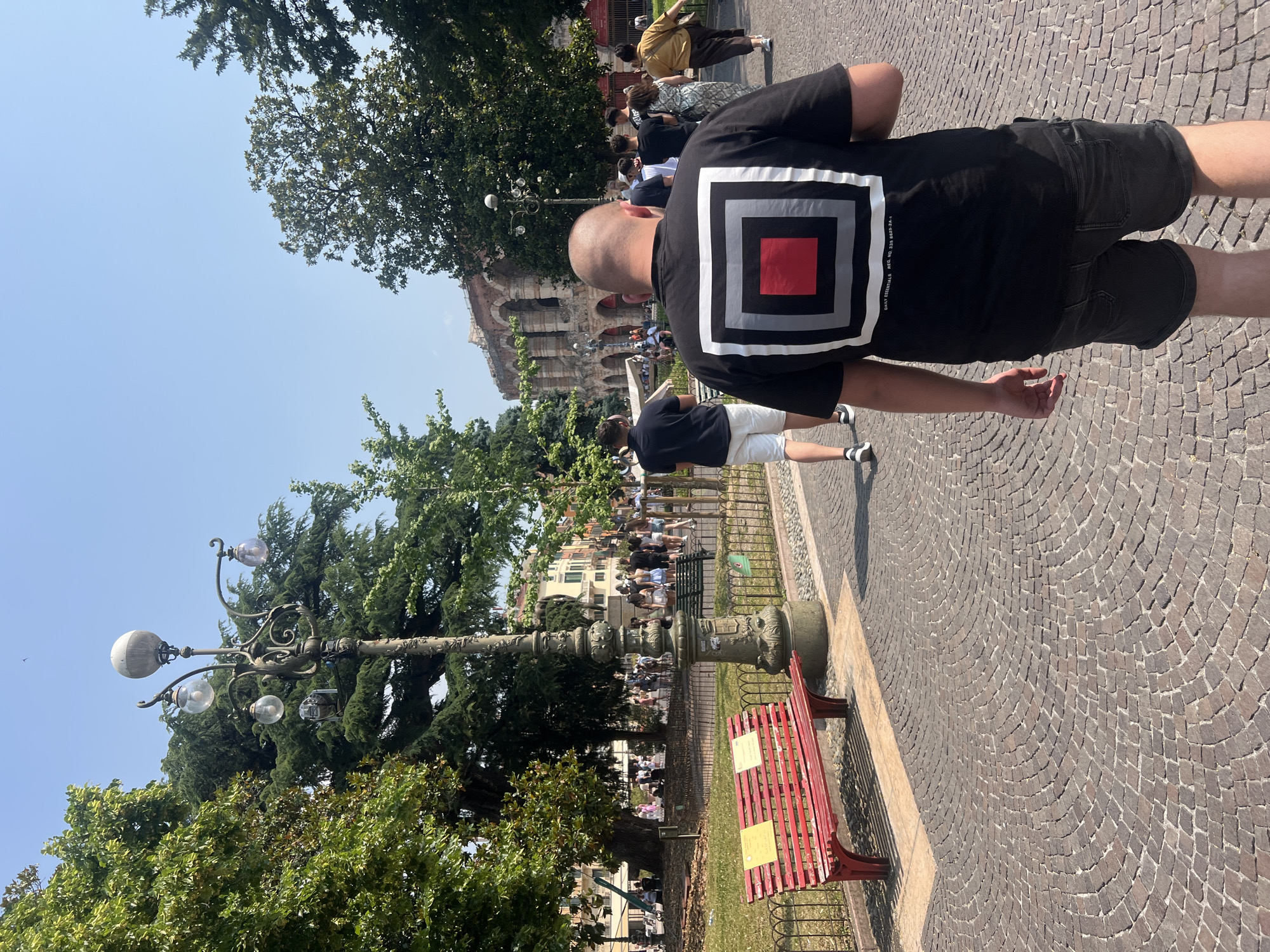 A low-angle outdoor shot depicts a bald man wearing a black T-shirt with a square graphic, facing away from the viewer. He is walking on a cobblestone path. To the left, there is an ornate lamppost and a red bench behind a low iron fence. In the background, the structure of a large colosseum-like building can be seen, along with a crowd of people. Trees are interspersed throughout the scene, and the sky is a clear blue. The location is the city of Verona, Italy, near the Verona Arena.