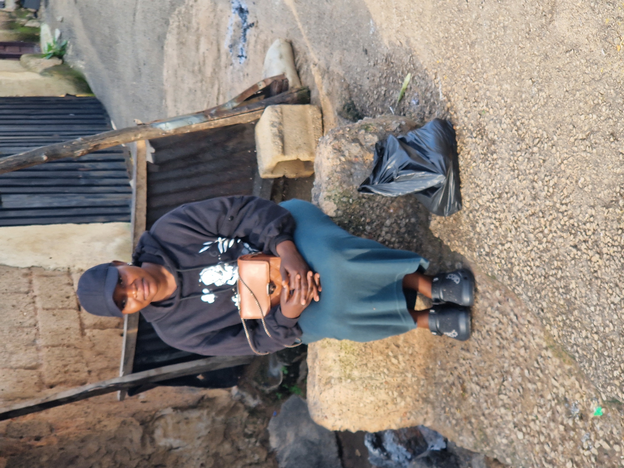 A young woman wearing a dark blue baseball cap, a black hoodie with a white graphic design, and a long, dark blue skirt sits on a rough, beige-colored rock. She holds a small, tan-colored purse with a gold chain strap in her lap, her hands clasped over it. She is wearing dark slip-on shoes with white embellishments. To her right, on the ground, is a black plastic bag. The background shows a rough, uneven wall constructed of dark bricks, and a wooden structure with corrugated metal sheeting. The ground is a mixture of dirt and small pebbles. The scene appears to be outdoors, in a somewhat unkempt urban setting, consistent with an area within the city of Jos, Nigeria. The lighting suggests it is daytime.