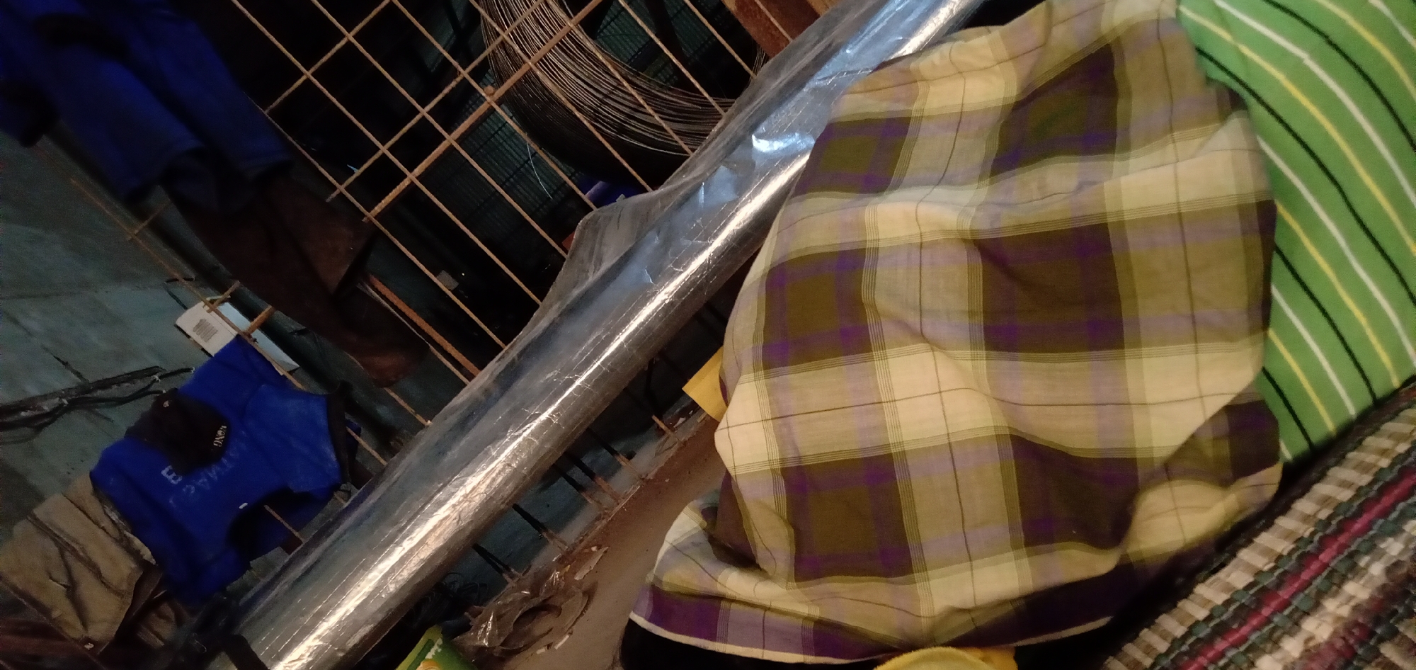 The image shows a cluttered indoor space. A roll of material wrapped in plastic leans diagonally from the lower left to the upper right of the frame. Behind this roll is a rusty wire mesh. To the left, there are clothes hanging, including a blue shirt with white lettering. To the right of the plastic-wrapped roll, there are various fabrics and blankets in different patterns, including plaid and stripes. The location is unknown.