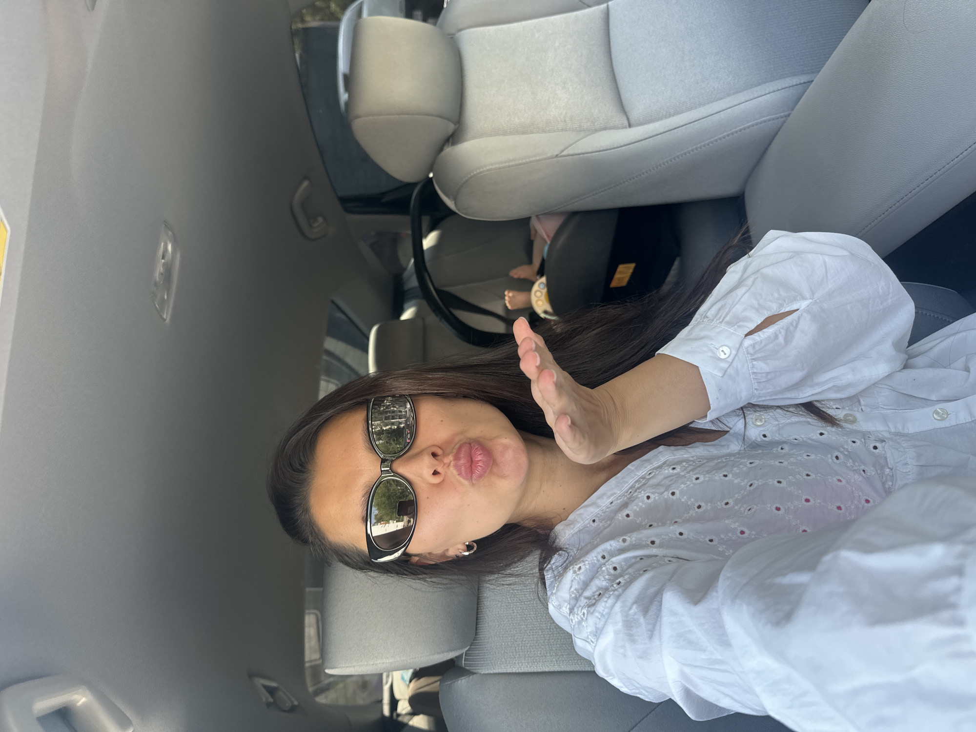 A woman in sunglasses and a white eyelet shirt sits in the driver's seat of a car, blowing a kiss. A baby car seat with a baby is visible in the back seat. The car interior is light gray.