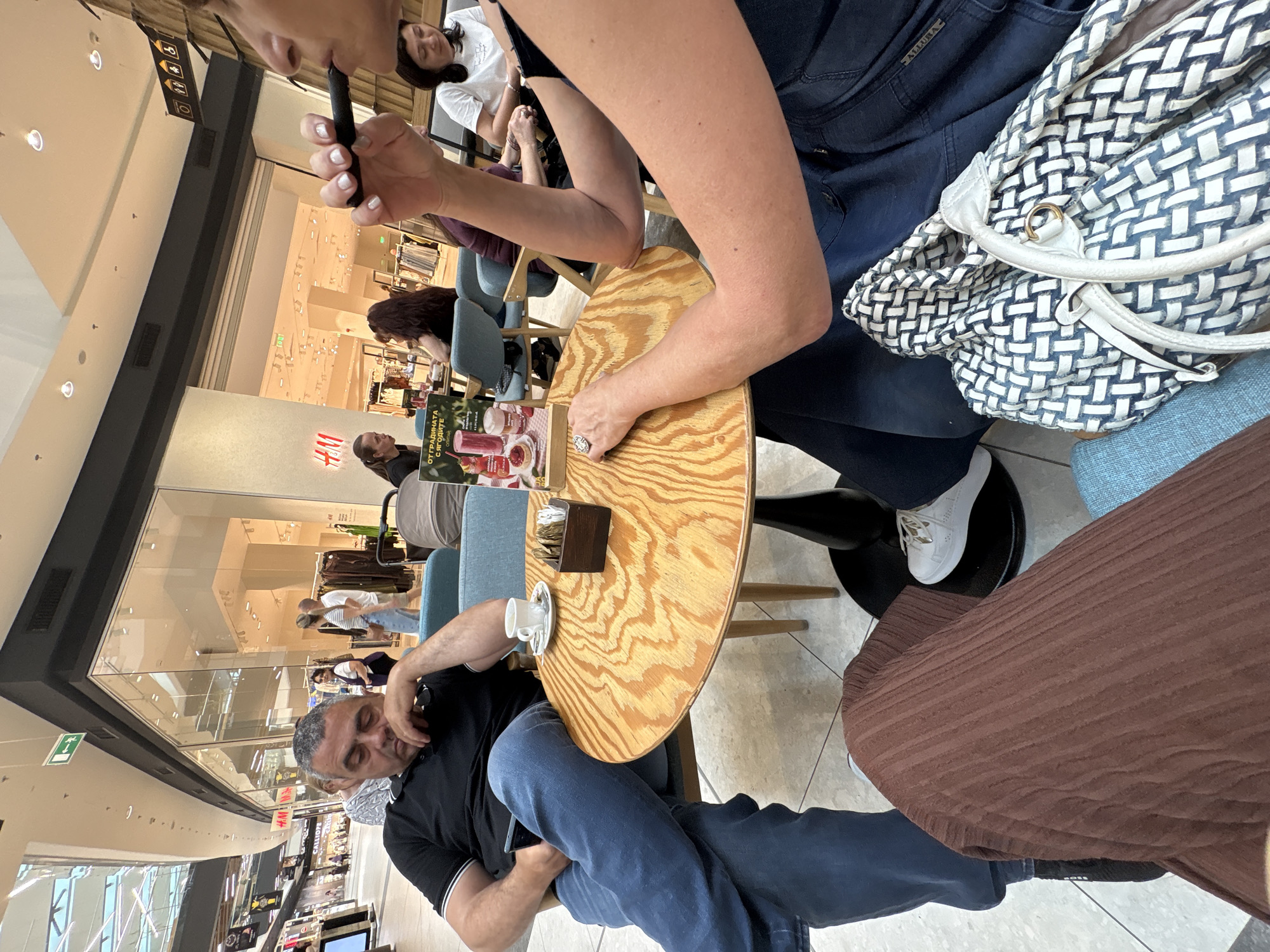 The image depicts an indoor scene, possibly a cafe or a restaurant within a shopping mall. A man is seated at a round wooden table. He wears a black shirt with white trim and blue jeans. His eyes are closed. A white ceramic cup and saucer are on the table near him. There is a wooden box next to the cup. At the table, an unidentified woman is wearing a navy shirt. Part of a woven white and blue bag is visible. A woman is smoking a black e-cigarette. The background shows clothing stores, indicating the location is a shopping mall. The H&M store sign is visible. Signs are in Bulgarian. Based on the signs and the location within a mall, it is plausible that the location is in Burgas, Bulgaria.
