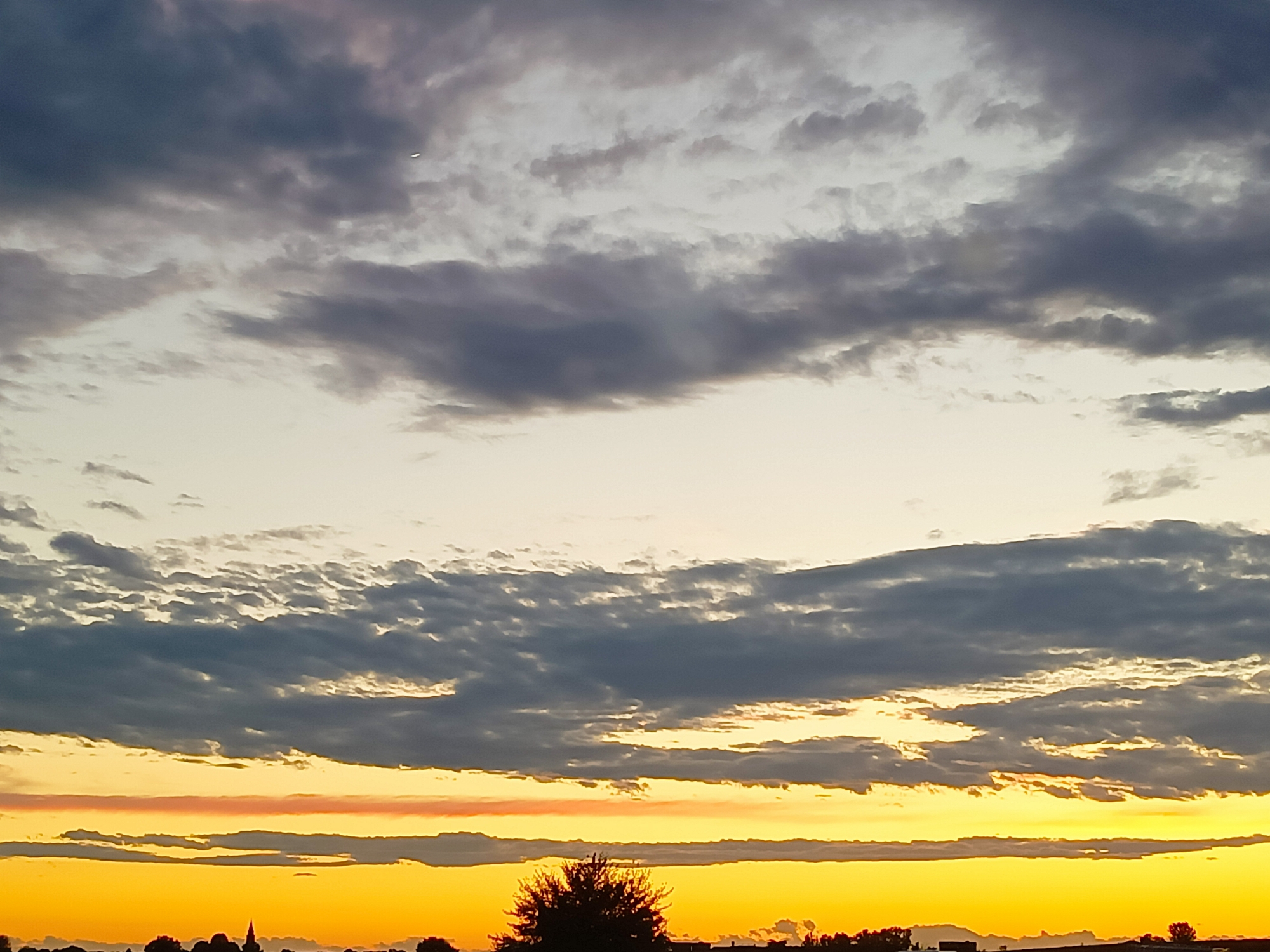 This is an outdoor photograph taken at sunset in Somaglia, Italy. The image captures a wide sky filled with dramatic clouds. A bright, intense yellow and orange glow emanates from the horizon, suggesting the sun is very low. Layered above this vibrant band are darker, greyish clouds, some of which are illuminated from beneath by the sunset's light. The upper portion of the sky transitions to a lighter, more neutral tone with wispy clouds. In the lower part of the frame, a silhouetted treeline stretches across the landscape, interrupted by the faint outlines of buildings and a prominent church spire. A single, small, dark object, possibly an airplane, is visible in the upper sky.