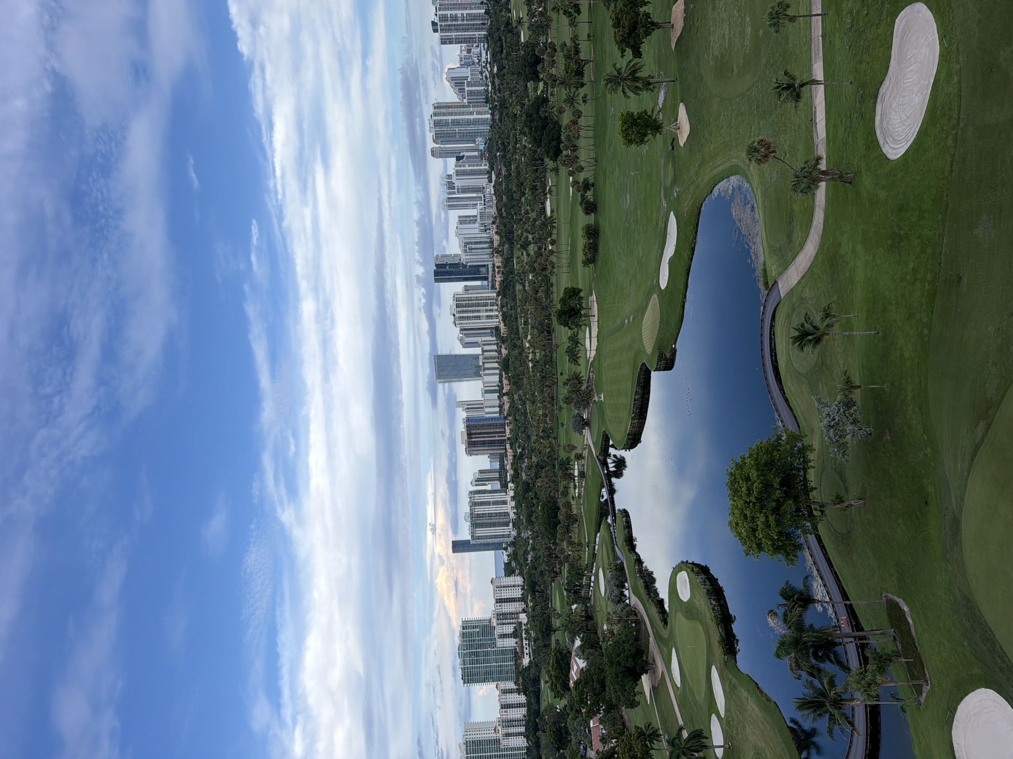 This aerial photograph captures a lush golf course adjacent to a cityscape in Aventura, Florida, USA. The foreground features a man-made lake with a dark, reflective surface, surrounded by well-maintained green fairways and sand traps. Several palm trees and other greenery are interspersed throughout the course. A winding pathway, possibly a cart path or pedestrian walkway, encircles parts of the lake. In the background, a dense cluster of high-rise buildings, indicative of a coastal urban environment, rises against a sky with scattered clouds. The buildings vary in height and architectural style, with some featuring balconies and glass facades. The overall scene depicts a harmonious blend of recreational space and urban development.