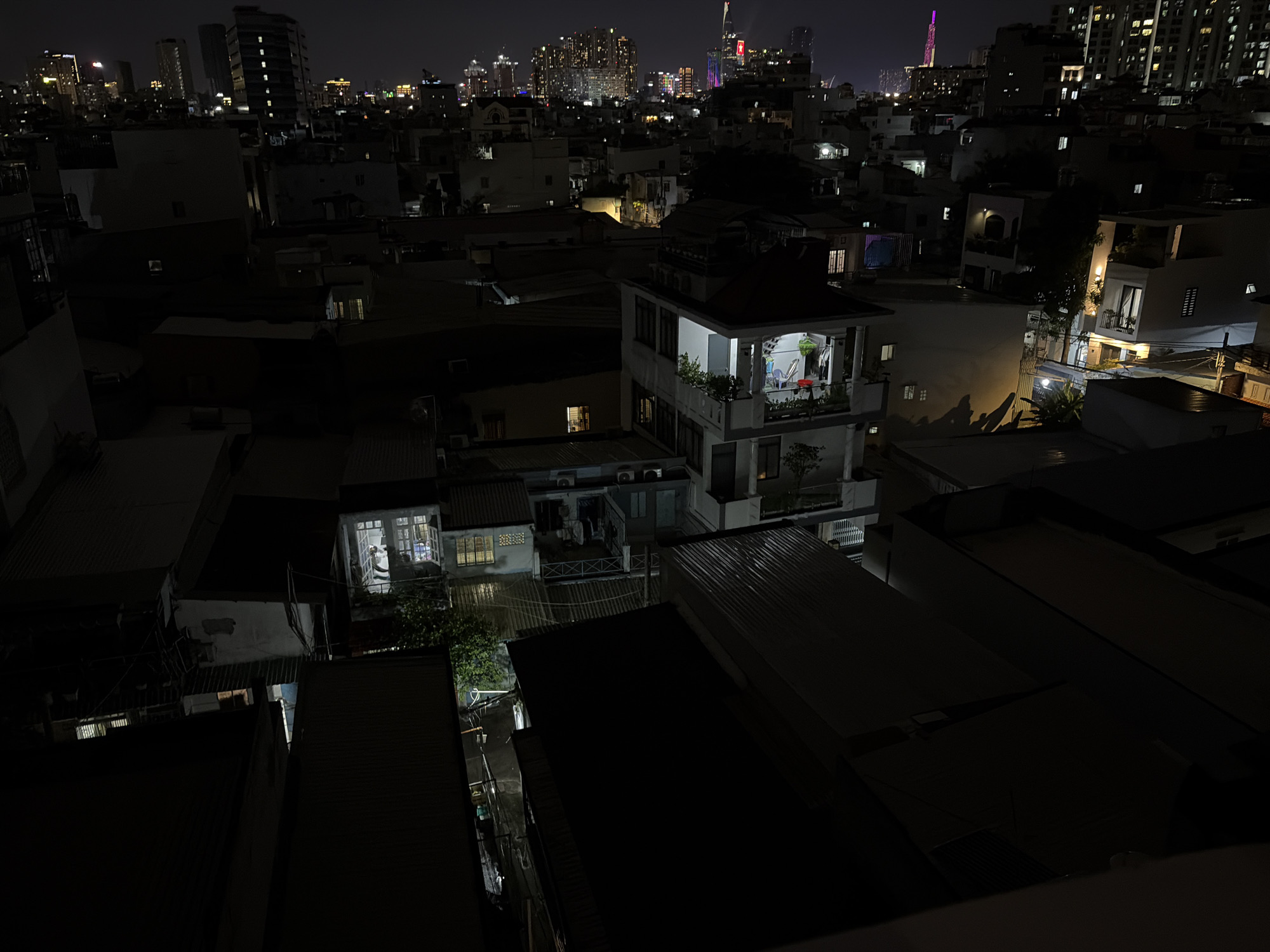 The image depicts a night scene in Thủ Đức, Vietnam, showcasing a dense urban environment characterized by a mix of low-rise buildings and a distant cityscape dominated by illuminated skyscrapers. The foreground is occupied by several dark, angled rooftops, suggesting a vantage point from a higher elevation. In the mid-ground, a distinctive multi-story building stands out with its brightly lit upper floors, revealing an interior with furniture and what appear to be people present. This building is surrounded by other structures, some of which also have illuminated windows, hinting at ongoing activity within. Further in the background, the metropolitan skyline is aglow with countless lights from taller buildings. The overall impression is one of a bustling city at night, with pockets of warm light emerging from residential and commercial spaces amidst the darkness.