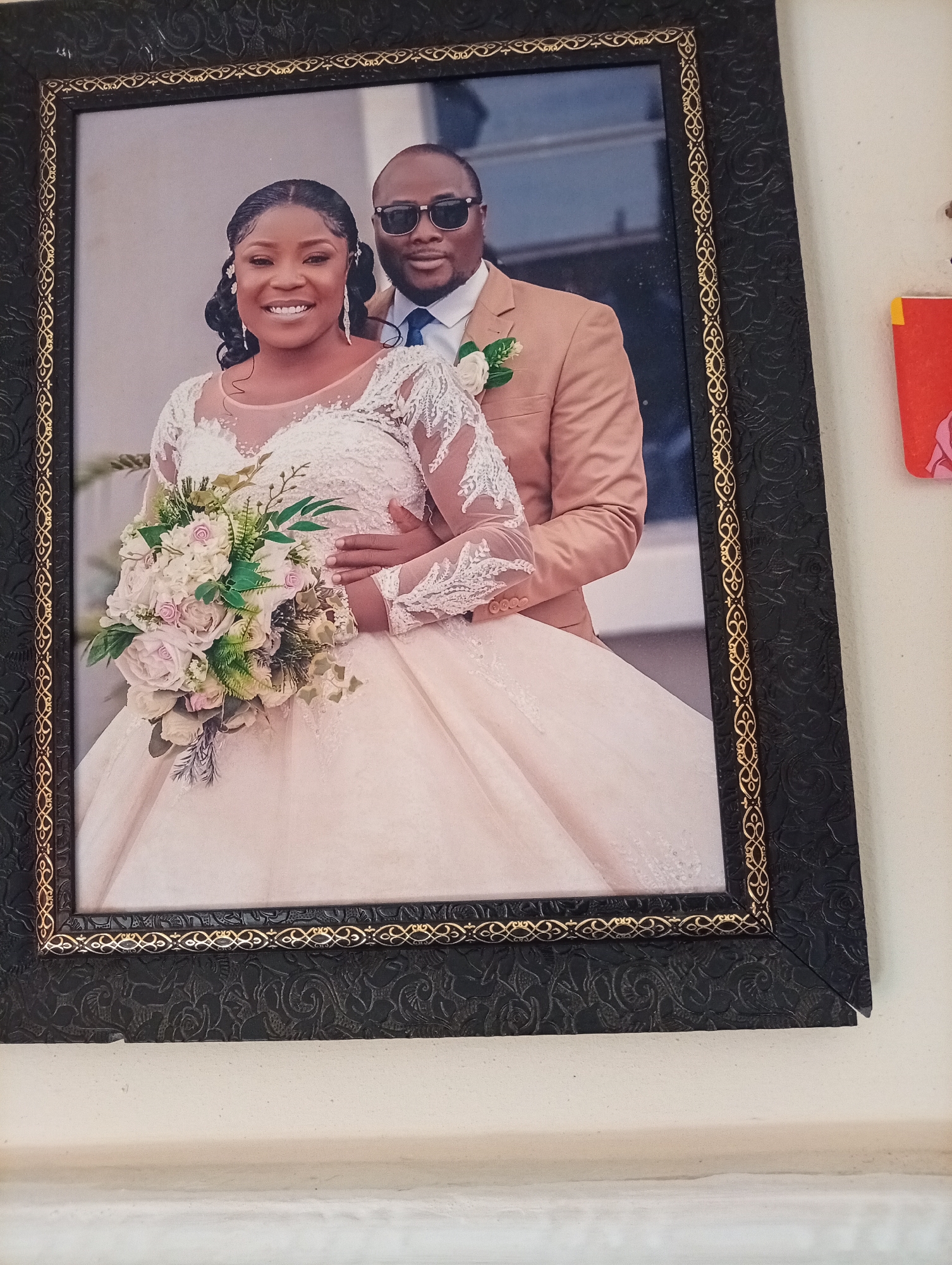 This is a framed wedding photograph of a Nigerian couple. The bride, on the left, wears a white wedding dress with intricate lace detailing on the sleeves and bodice. She holds a bouquet of light-colored roses and greenery. The groom, on the right, is dressed in a tan suit with a blue tie and sunglasses. They are both smiling and looking towards the camera. The photograph is in a black frame with a decorative gold trim. The background of the photograph appears to be an outdoor setting with a building. The photograph is hanging on a white wall. This image was taken in Gura Top, Nigeria.