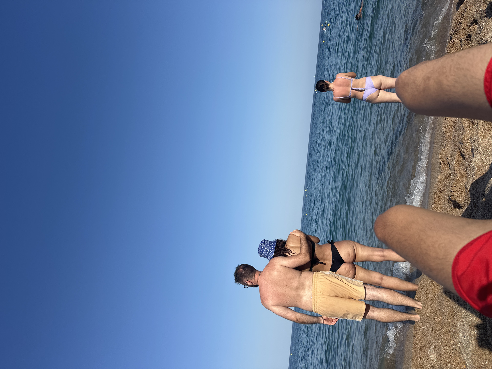 In Blanes, Spain, on a clear, sunny day, three individuals are seen on a sandy beach. In the foreground, a person's legs, clad in red swim trunks, are positioned prominently, suggesting the viewpoint of someone lying down. Further back, a couple stands near the water's edge. The man, facing away from the camera, wears light brown swim trunks and glasses. He has his arm around the woman beside him, who is wearing a black bikini and a blue patterned bucket hat. She is also facing away from the camera. In the distance, a third woman stands in the shallow water, facing away from the camera. She is wearing a light purple bikini. The ocean is calm and blue, with yellow buoys visible in the water. The sky is a vibrant, clear blue.