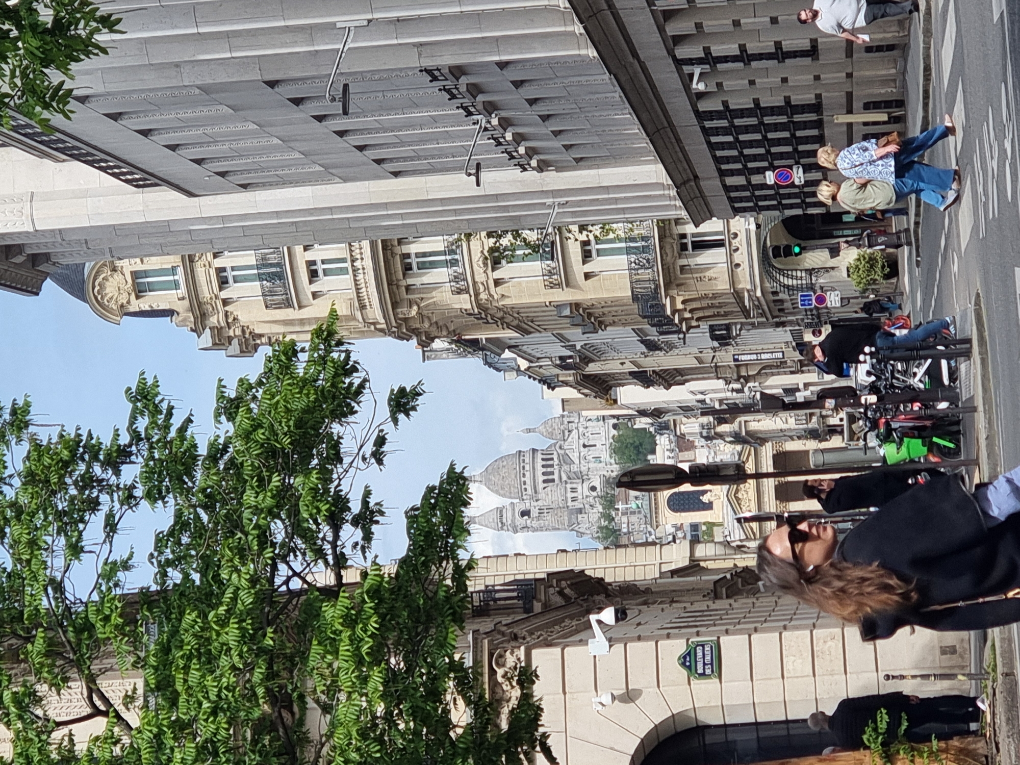 The image captures a vibrant daytime street scene in Paris, France, with the iconic Sacré-Cœur Basilica majestically visible in the distance. The perspective looks down a wide boulevard lined with classic multi-story Haussmannian buildings, characterized by their light stone facades, ornate balconies, and detailed architectural elements.
In the immediate foreground, a lush green tree with dense foliage partially frames the left side of the scene. On the right, several pedestrians animate the street. A woman with long brown hair and sunglasses is prominently featured in the mid-ground, looking upwards. Further down, two individuals walk on the sidewalk, and another person stands near the far end of the street. Bicycles are parked or being ridden, and a green traffic light is visible, indicating an active street.
Notable details include several security cameras mounted on the buildings. A distinct green street sign on the right building clearly reads "BOULEVARD DES ITALIENS," confirming the Parisian location. Another red sign with partially obscured text appears to say "FOOD & BAGUETTE" or similar. The clear sky and strong shadows suggest bright daylight, likely mid-morning or afternoon. The overall atmosphere conveys a typical busy Parisian street.