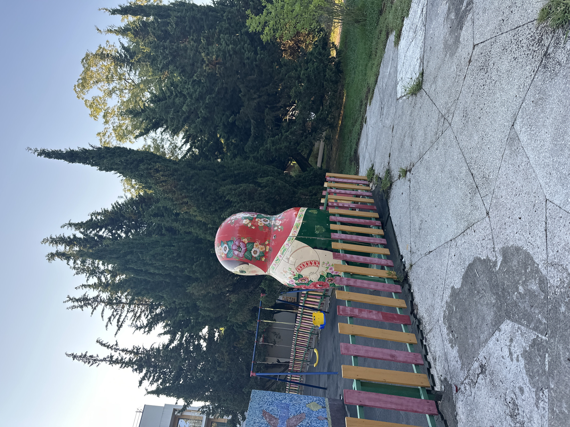 The image depicts a children's playground located in Sochi, Russia, characterized by a large, intricately painted Matryoshka doll standing prominently near a colorful wooden fence. The playground features swings, a slide, and a mosaic wall. The environment is lush with tall cypress trees and other greenery, set against a clear blue sky. The concrete ground shows signs of wear, with cracks and a damp patch. The scene is tranquil, with no people present.