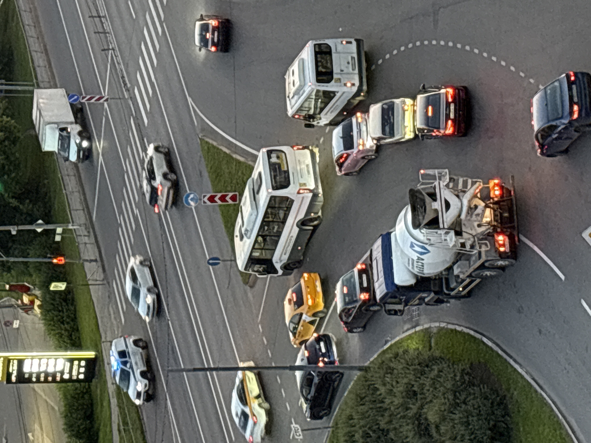 The image shows a traffic scene in a city, likely during the day or dusk. Several vehicles are visible on a multi-lane road and an intersection.
In the foreground, a cement mixer truck with a blue cab is driving on a curved road. To its right, a dark-colored sedan is following. Further up the road, a light-colored car is in front of another dark-colored sedan. There are also two white mini-buses, one to the right of a pink car, and another bus slightly further back and to the left. A yellow taxi is turning at the intersection.
In the middle ground, a white passenger bus is driving on the main road. A white car is to its left. Further up, a white SUV is driving on the road.
In the background, a white truck is driving on the road. A white police car with flashing blue lights is visible on the left side of the image, stopped or moving slowly. A white sedan is behind the police car.
The environment appears to be urban, with a traffic light visible in the upper left corner, indicating a controlled intersection. There are also pedestrian crossings marked on the road. Green vegetation lines the sides of the road in some areas.