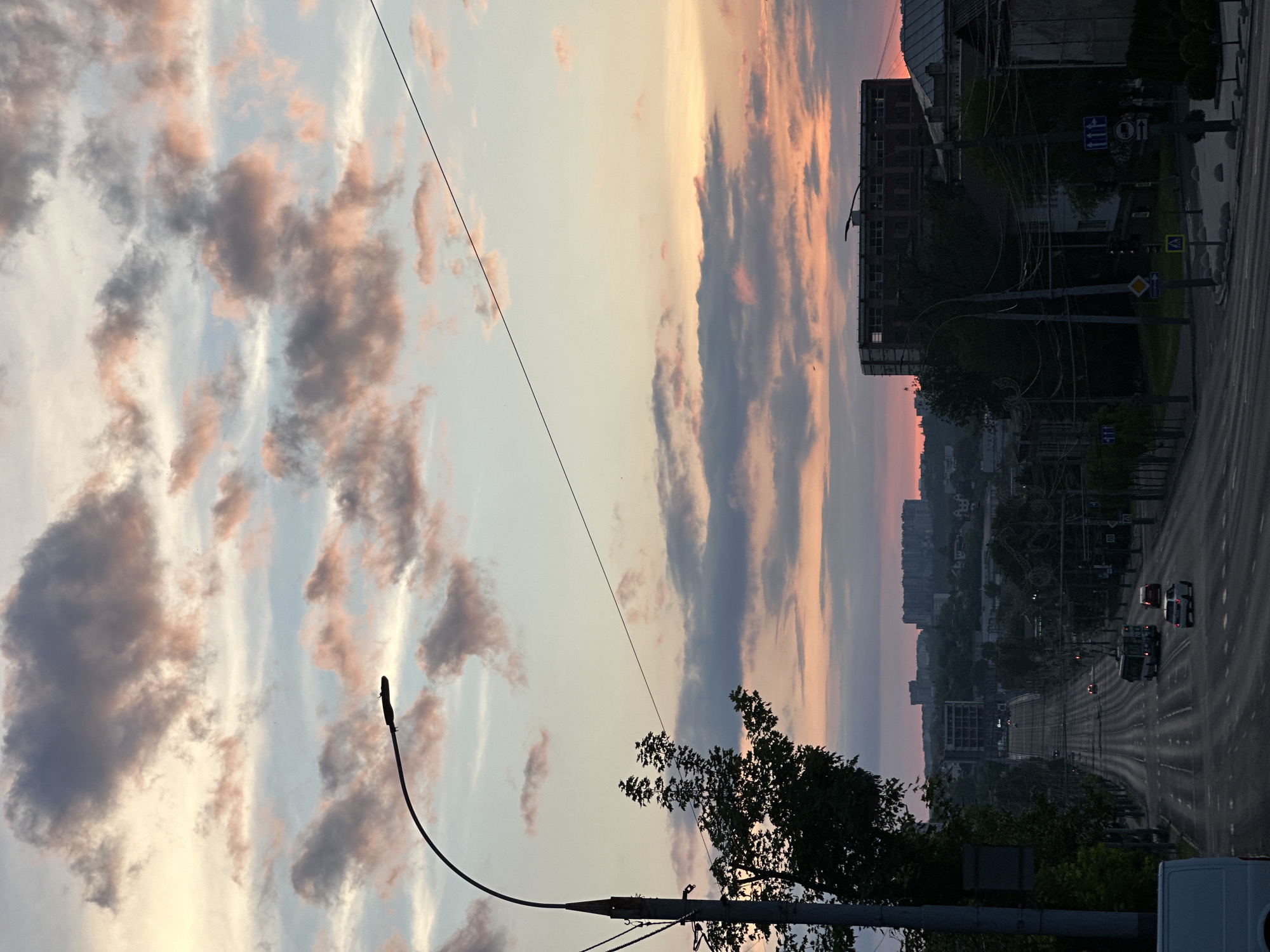 The image shows a street scene at sunset in Chișinău, Moldova. The sky is filled with pink and gray clouds. A street lamp is visible in the foreground, along with tree silhouettes. The road stretches into the distance with several cars on the road. Buildings and trees are visible along the sides of the street. Road signs are present. An unfinished building is on the left side of the road. The lighting suggests it's dusk.