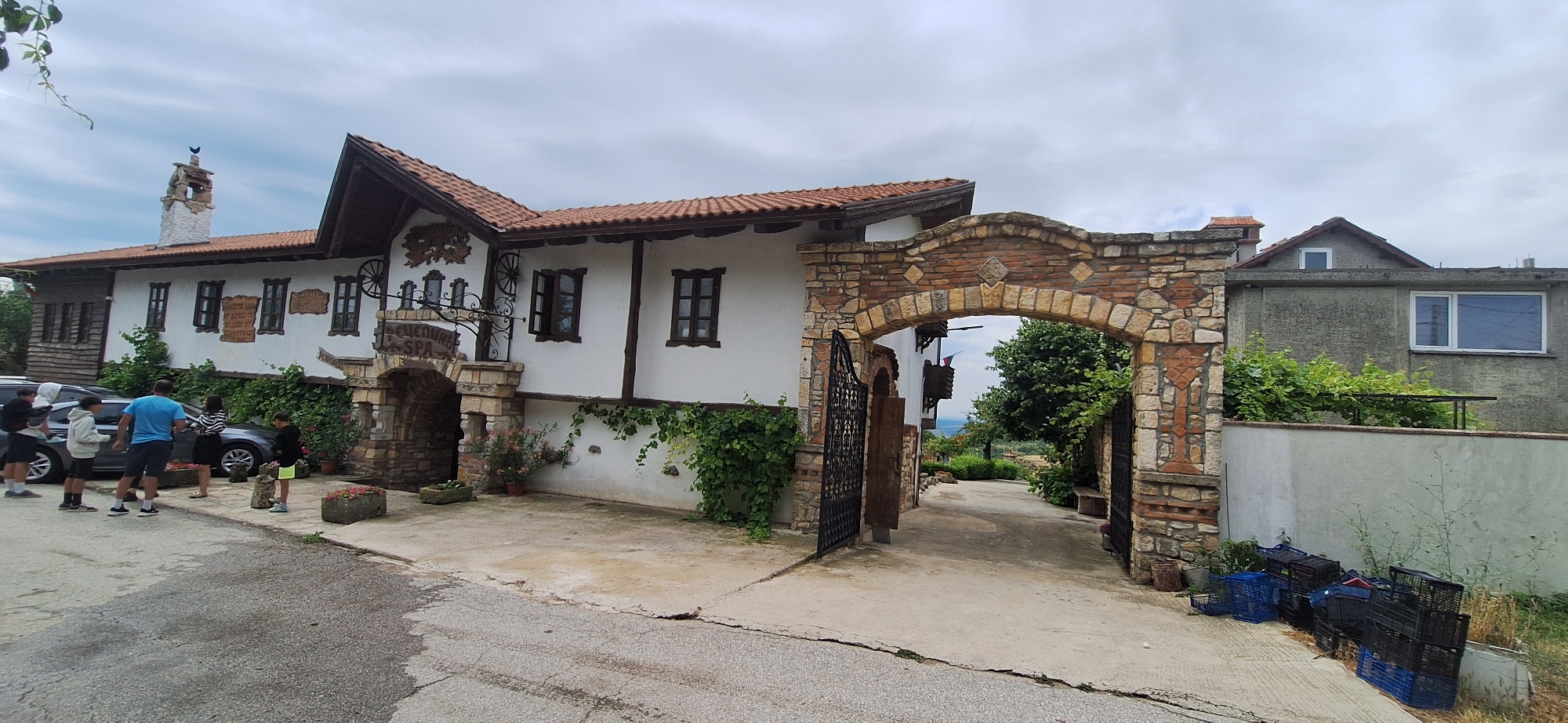 The image shows the entrance to a traditional Bulgarian guesthouse and spa complex in Kalimantsi, Bulgaria. The entrance features a rustic stone archway with metal gates and is flanked by a white building with a red tile roof. The building has decorative wooden elements and several windows. In front of the building, several people are standing near a car. A paved driveway leads through the archway, revealing a glimpse of greenery and a distant view. To the right of the archway, a more modern, grey building is visible, with some plastic crates piled near its base. The sky is overcast, suggesting a cloudy day.