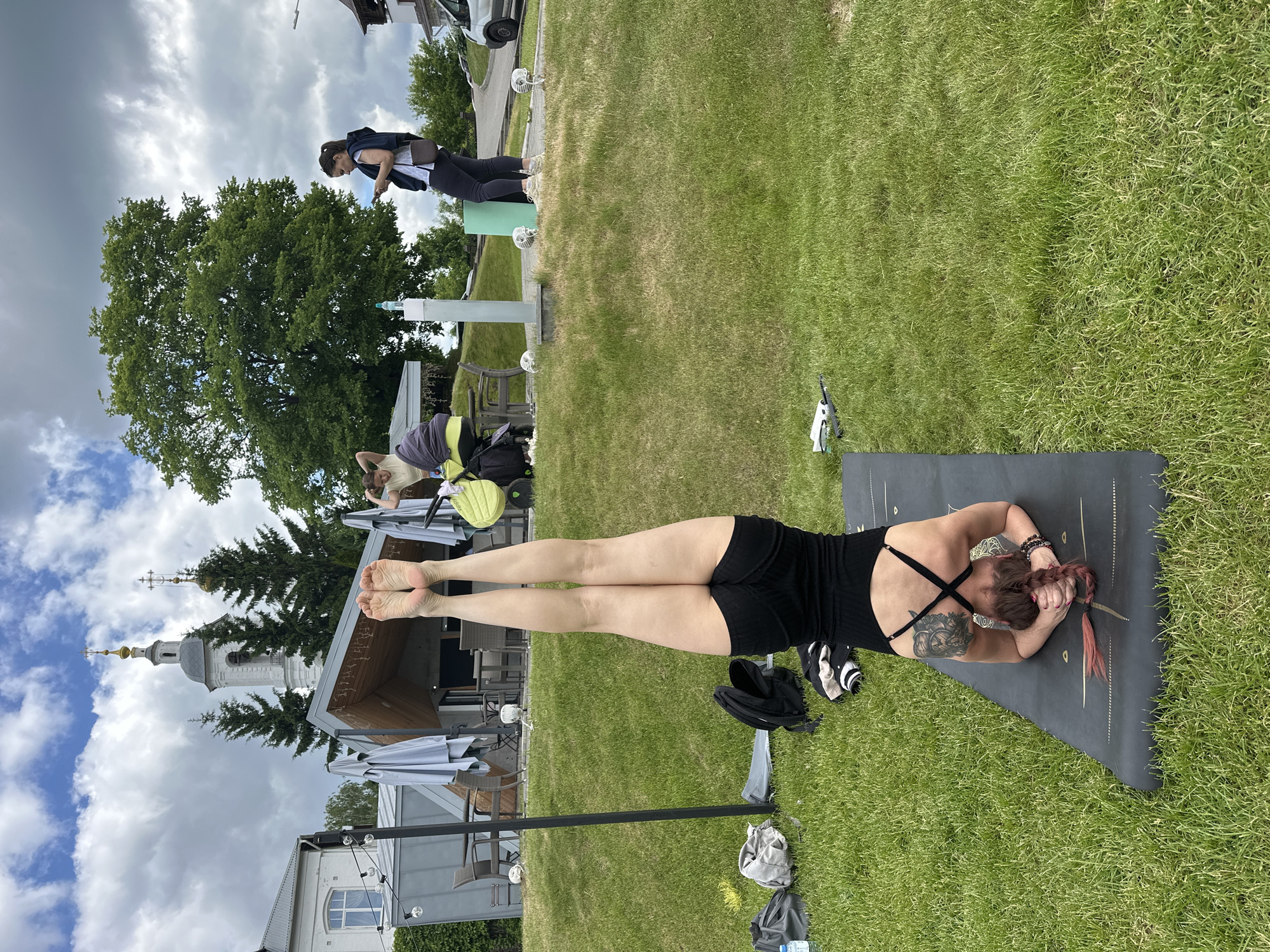 An outdoor scene captures a woman performing a yoga pose on a dark gray mat laid out on green grass. She is lying prone, dressed in a black tank top and shorts, with her bare feet raised vertically towards the sky. Her head is turned slightly to the right, and her hands are clasped under her chin. A tattoo of a mythical creature is visible on her upper back/shoulder area, and her reddish-brown hair, with hints of pink, is in a braid. Next to her mat are black sneakers, a gray backpack, and another small bag.
In the mid-ground, other people are present on a grassy area surrounded by outdoor furniture, including tables and umbrellas. One woman stands on a light green platform, looking down, dressed in a dark jacket and pants. Another woman is seated near a yellow and gray stroller or large bag.
The background features lush green trees and traditional architecture, notably a white church steeple with an onion dome and a golden cross, characteristic of Russian Orthodox churches. Other buildings are partially visible. The sky is overcast with fluffy white and gray clouds. The overall setting appears to be a park or an outdoor relaxation area, possibly connected to a café or restaurant, in Vladimir, Russia. The scene depicts daytime under a cloudy sky. No discernible text is visible in the image.