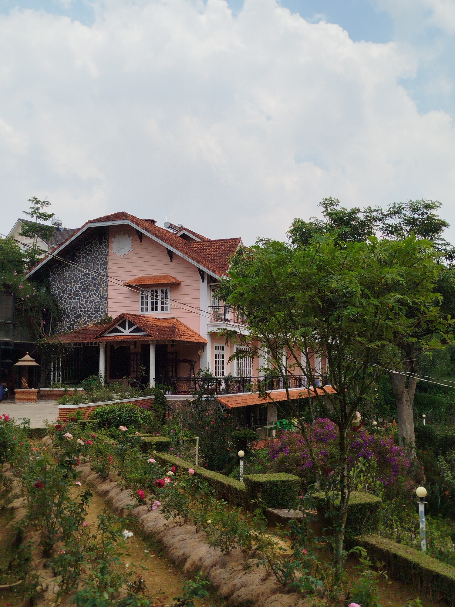 The image depicts a two-story building with a pink facade and a red-tiled roof, featuring a stone section on one side. It is situated on a hillside with a terraced garden. The garden contains rows of roses and manicured hedges. A large tree is visible on the right side of the frame. The sky is partly cloudy. The building appears to be a villa or guesthouse, based on its architecture and setting. The location is likely Lang Biang - Đà Lạt, Vietnam, given the building style and the vegetation. There are no people visible in the main frame. The foreground consists of the terraced garden.