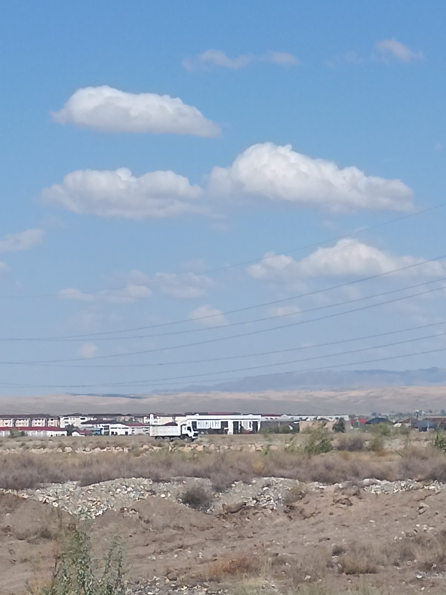 A wide shot captures a suburban landscape in Taldykorgan, Kazakhstan. A clear blue sky with scattered white clouds dominates the upper half of the image. In the middle ground, a row of buildings, some with red roofs and others with white facades, stretches horizontally. A white truck with a large container is parked in front of one of these buildings. Utility lines are visible against the sky. The foreground consists of dry, scrubby vegetation and rocky terrain, suggesting an arid environment. In the far background, a range of low, hazy hills forms the horizon.