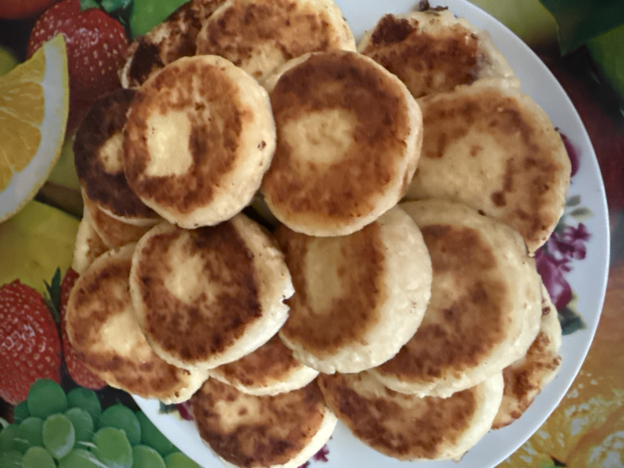 A plate of golden-brown, circular syrniki, a type of cheese pancake, is piled high on a white plate. The syrniki have a slightly browned exterior and a lighter, fluffy interior. The plate is placed on a colorful tablecloth with a fruit pattern, featuring images of strawberries, grapes, and citrus slices. The lighting is bright, suggesting an indoor setting, possibly a kitchen or dining area. There are no people or animals visible in the image. The scene is set in Sovietskyi, Ukraine.
