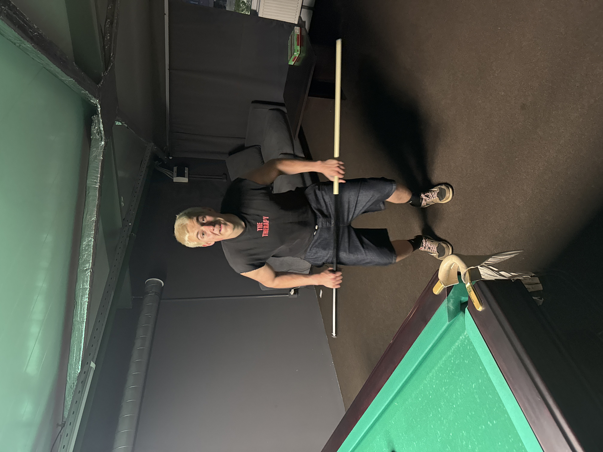 A young man with bleached blonde hair, wearing a black t-shirt with red text and denim shorts, is holding a pool cue in a dimly lit room. The room has dark gray walls and a dark ceiling with exposed pipes and insulation. To the left, a green pool table is visible, and to the right, a dark sofa and a coffee table are present. The man appears to be in the middle of a pool game, holding the cue in a ready position.