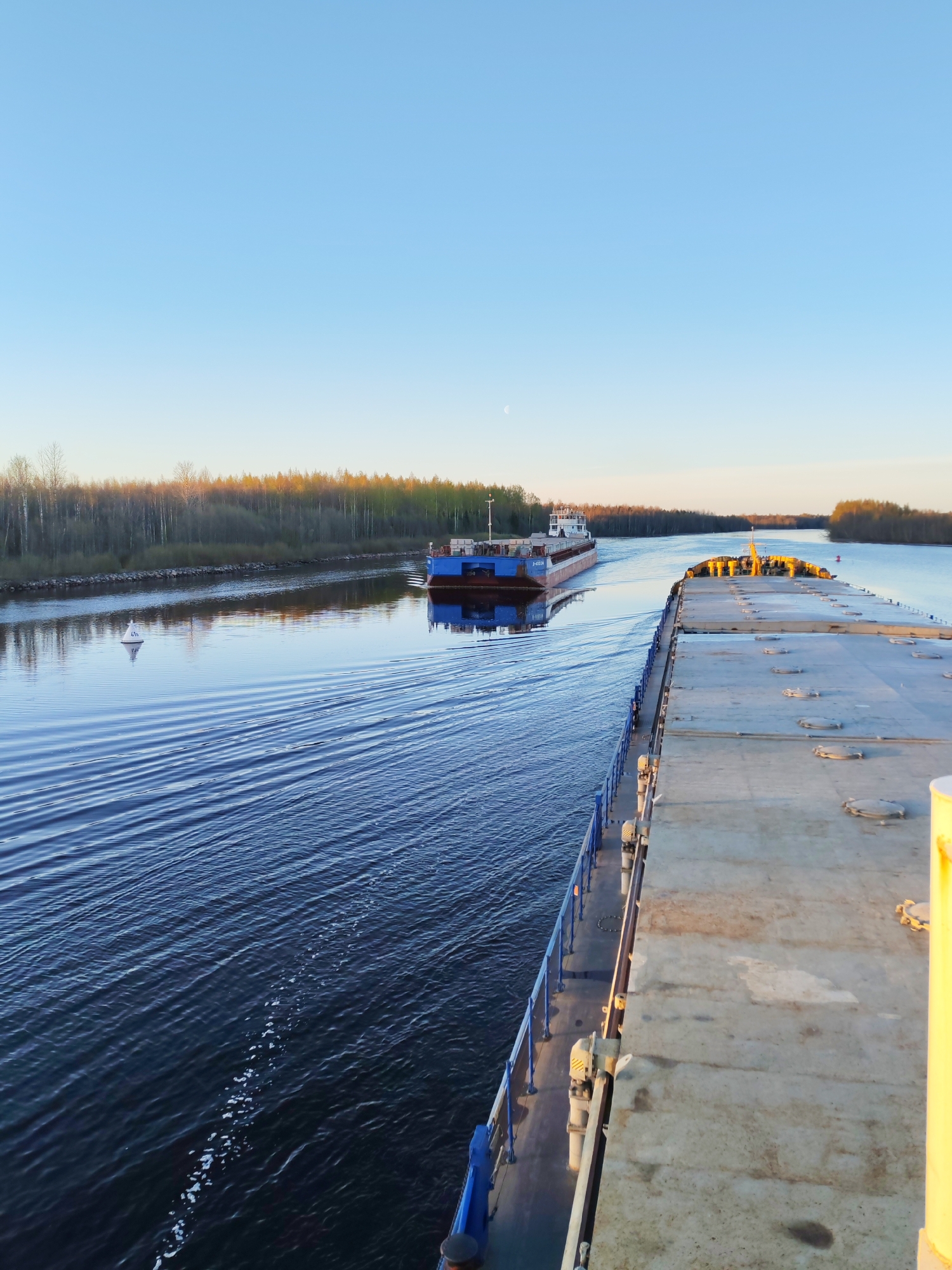A long shot captures two barges moving along a river near Kurdiug, Russia. The barge closest to the viewer is gray and occupies a significant portion of the right side of the frame. The other barge is a darker blue and is further away, moving in the opposite direction. Both vessels leave wakes in the water. The river reflects the clear blue sky. Trees line the riverbanks, with autumn colors beginning to appear on some of the leaves. A small, white buoy is visible in the river near the left bank. The time of day appears to be late afternoon.