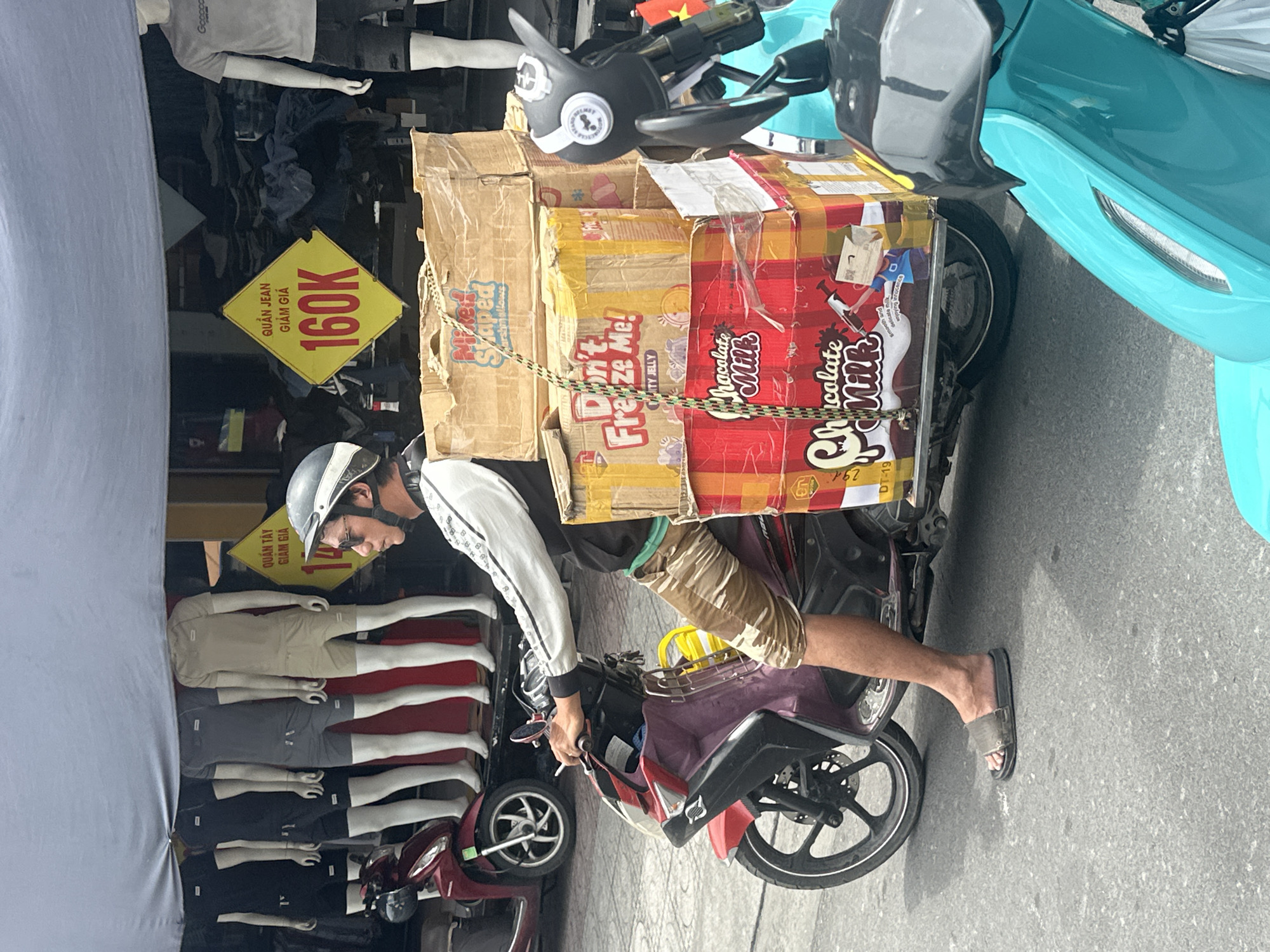 A man is seen on a purple and red scooter with a large stack of boxed goods tied to the back. The boxed goods include "Milk Shaped Marshmallows", "Don't Freeze Me! Fruity Jelly," and "Chocolate Milk". He is wearing a white jacket, khaki shorts, a gray helmet, and sandals. The background shows a storefront with mannequin legs displaying shorts and a sign for 160K quần jean. The photo appears to be taken in Bac Nha Trang Ward, Vietnam. The boxes stacked and strapped to the scooter are labelled with, "291 DT-19" and "P62" in the corner. Another scooter is partially visible to the left. A pale blue scooter is visible in the upper right corner.