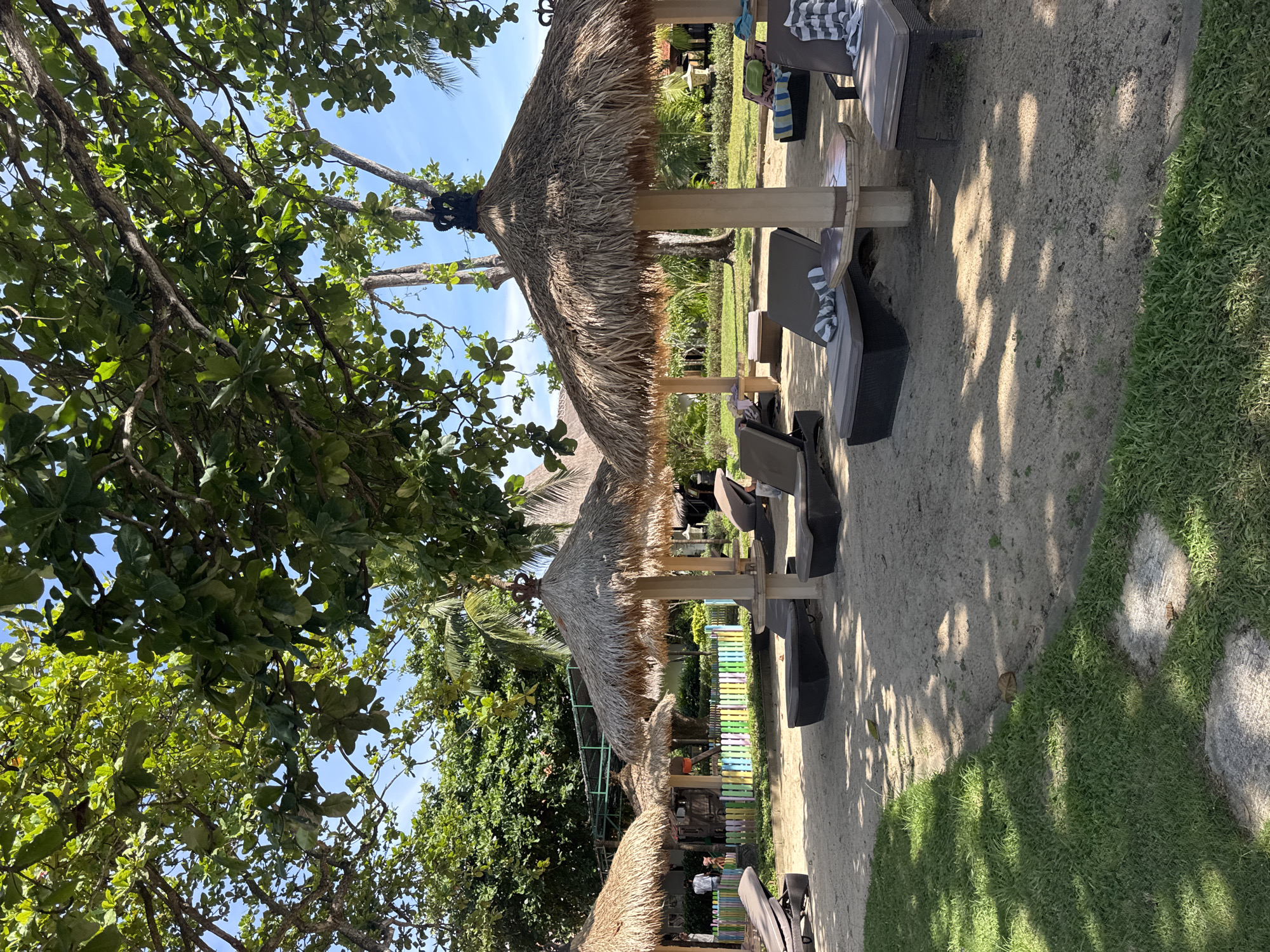 The image shows a beach resort scene in Kuta Selatan, Indonesia. There are four thatched-roof parasols providing shade over lounge chairs on a sandy area. A variety of green trees and plants surround the area. Some chairs have towels on them. A colorful fence can be seen in the background between the parasols.