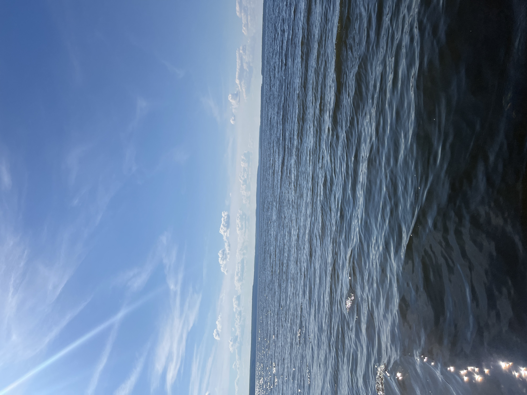 A serene view of a large body of water, likely a lake or river, under a bright blue sky with scattered white clouds. The sun's rays streak across the upper left portion of the sky, creating a luminous effect. The water's surface is rippled, reflecting the sunlight in shimmering patches, particularly in the foreground and mid-ground. In the distance, a long, low-lying shoreline is visible, suggesting a vast expanse of water. The image captures a tranquil, natural environment, without any visible people or man-made structures. The location is identified as Siukterka, Russia.
