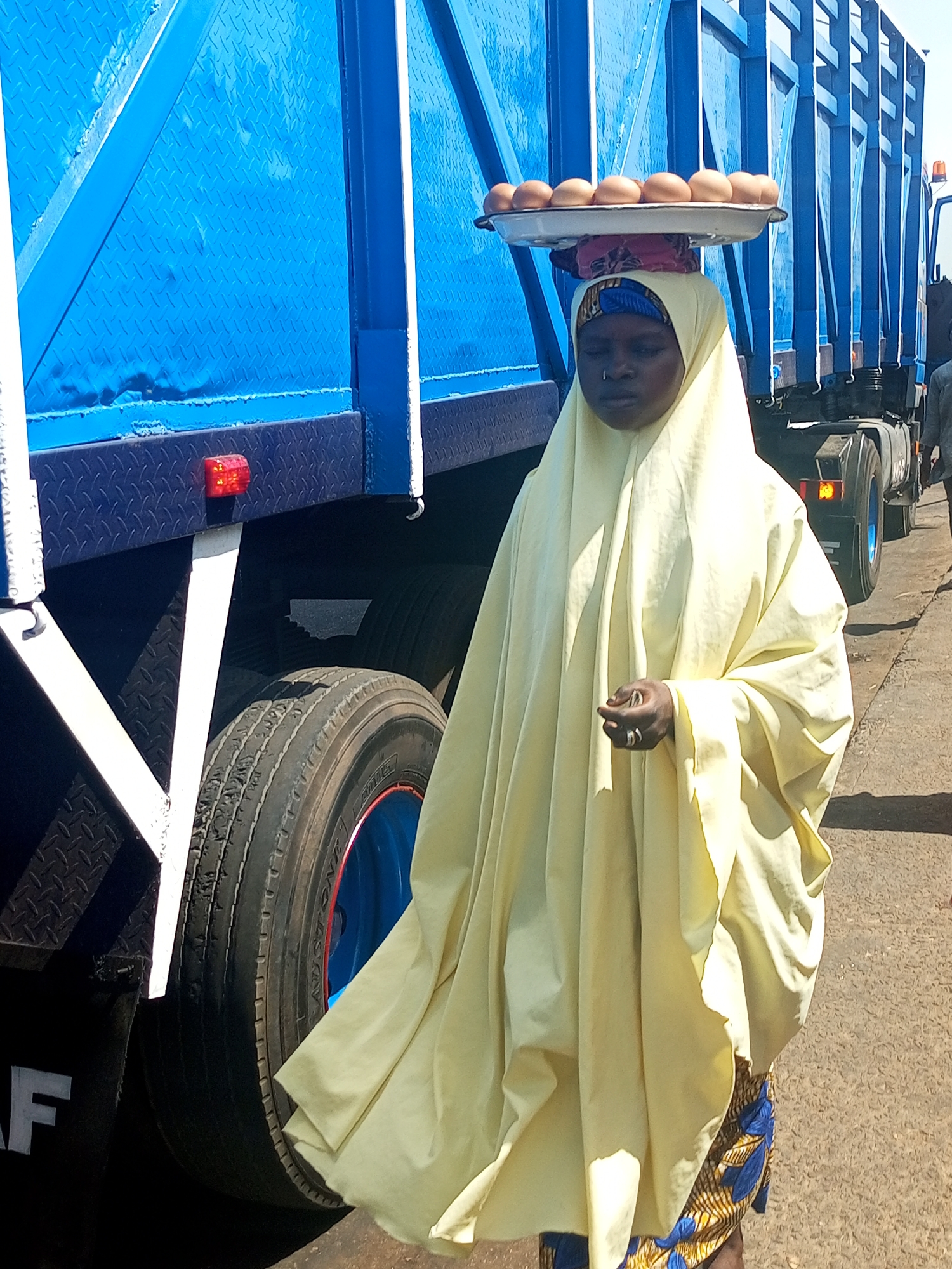 The image shows a person, likely a woman, in Gadao, Nigeria, standing next to a large blue commercial truck under bright daylight. She is carrying a flat white tray with approximately ten light brown eggs balanced on her head. She wears a flowing, light yellow garment that covers her body, along with a patterned headscarf underneath the tray. Her face is visible but appears serene or focused.
The dominant background is the side of the bright blue truck, which features a diamond plate texture on its metallic surface. A red rectangular reflector light is visible on the lower section of the truck's body. Below the truck, a large tire with a blue and red rim is prominent, showing wear. A black mudguard with the white letter "F" (likely part of the brand "DAF") is also visible. The ground appears to be a paved or hard-packed surface. The strong sunlight casts clear shadows, indicating it is daytime. The scene depicts a common sight of roadside vending or transport in the region.