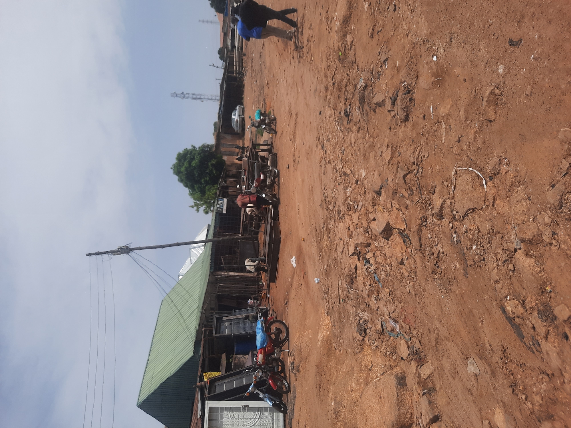 The image depicts a street in Gura Top, Nigeria. The ground is primarily reddish-brown dirt, scattered with rocks and debris. Several buildings line the street, one with a green metal roof. Power lines are visible. Parked motorcycles are present. Two individuals are walking in the background. A silver car is also parked further down the street.