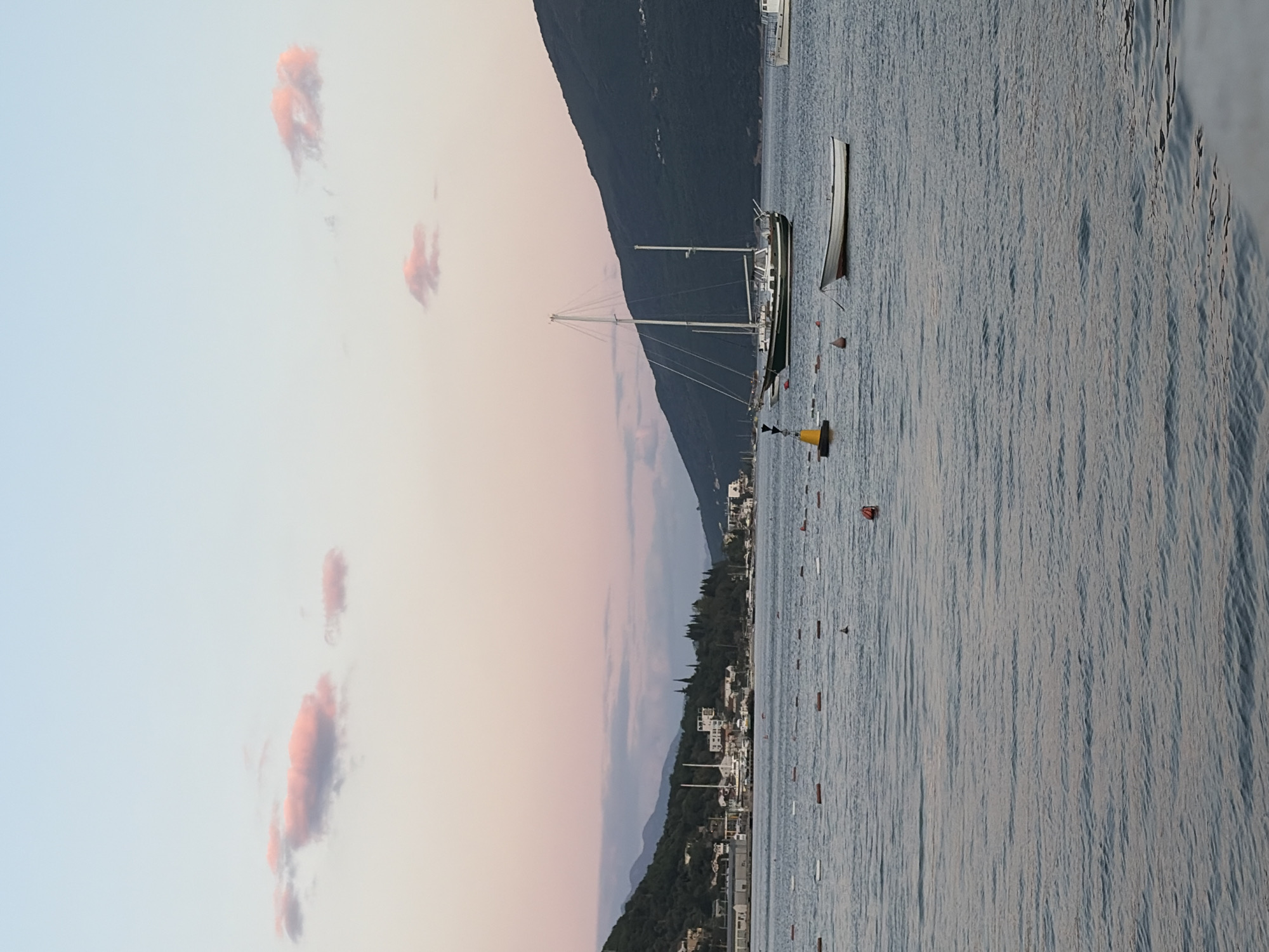 The scene depicts a coastal view in Meljine, Montenegro, featuring a sailboat anchored in calm, light blue water. A small rowboat is nearby. In the background is a mountainous terrain dotted with houses, receding into the horizon. A pink-hued sky contains scattered clouds. Buoys are on the water's surface.